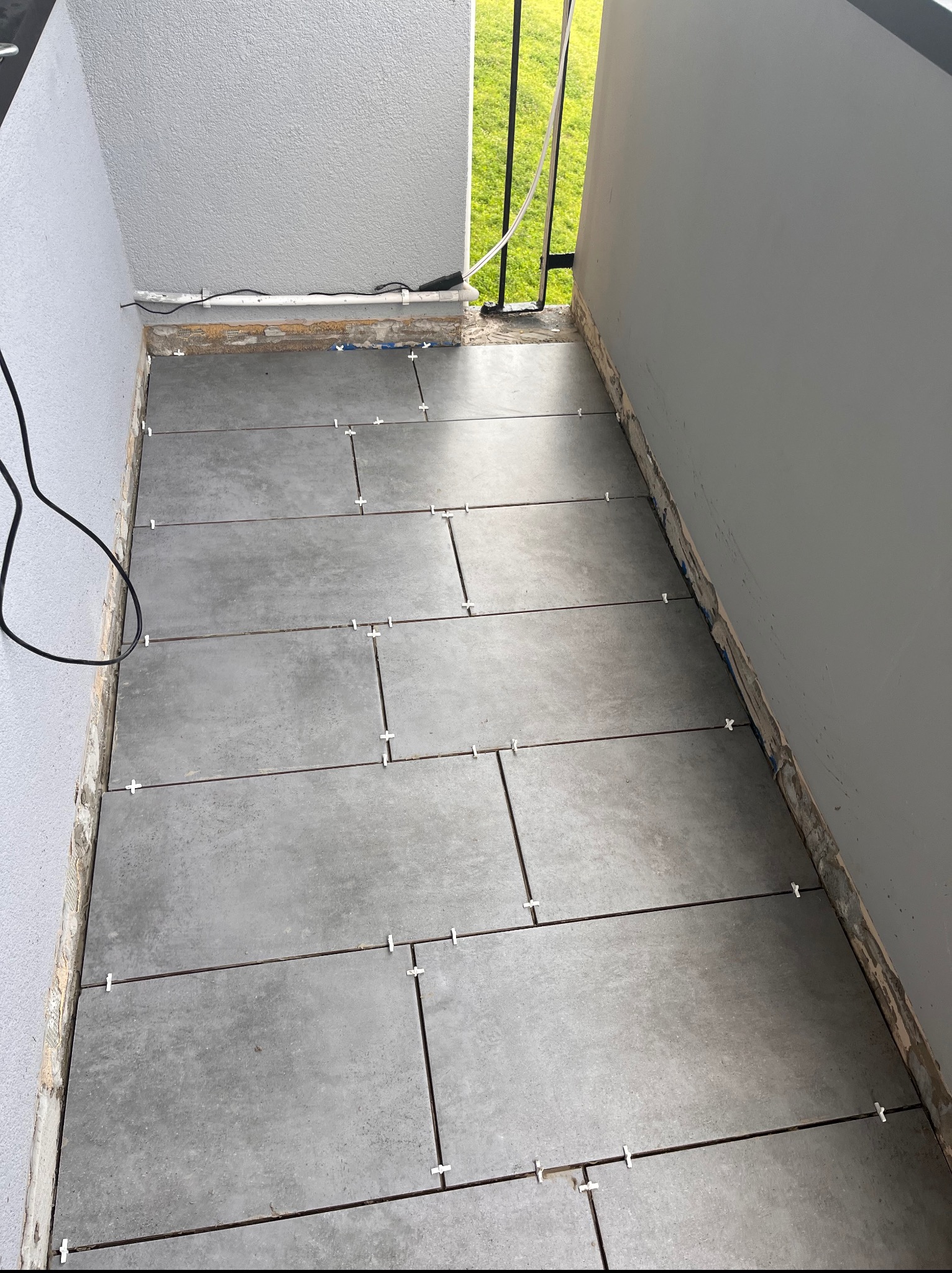 Układanie szarych płytek na balkonie z użyciem krzyżyków dystansowych. Widoczne kable i fragment zieleni w tle. Etap prac wykończeniowych na zewnątrz.