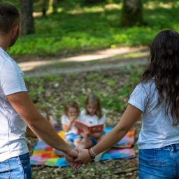 Olender Fotografia - Para trzymająca się za ręce, z tyłu dwie dziewczynki czytają książkę na kolorowym kocu w lesie. Ujęcie zza pleców, naturalne światło.