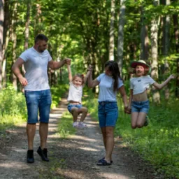 Uśmiechnięta rodzina na leśnej ścieżce: tata podnosi córkę, mama trzyma ją za rękę, a druga córka skacze. Wszyscy w letnich ubraniach, na tle zieleni drzew.