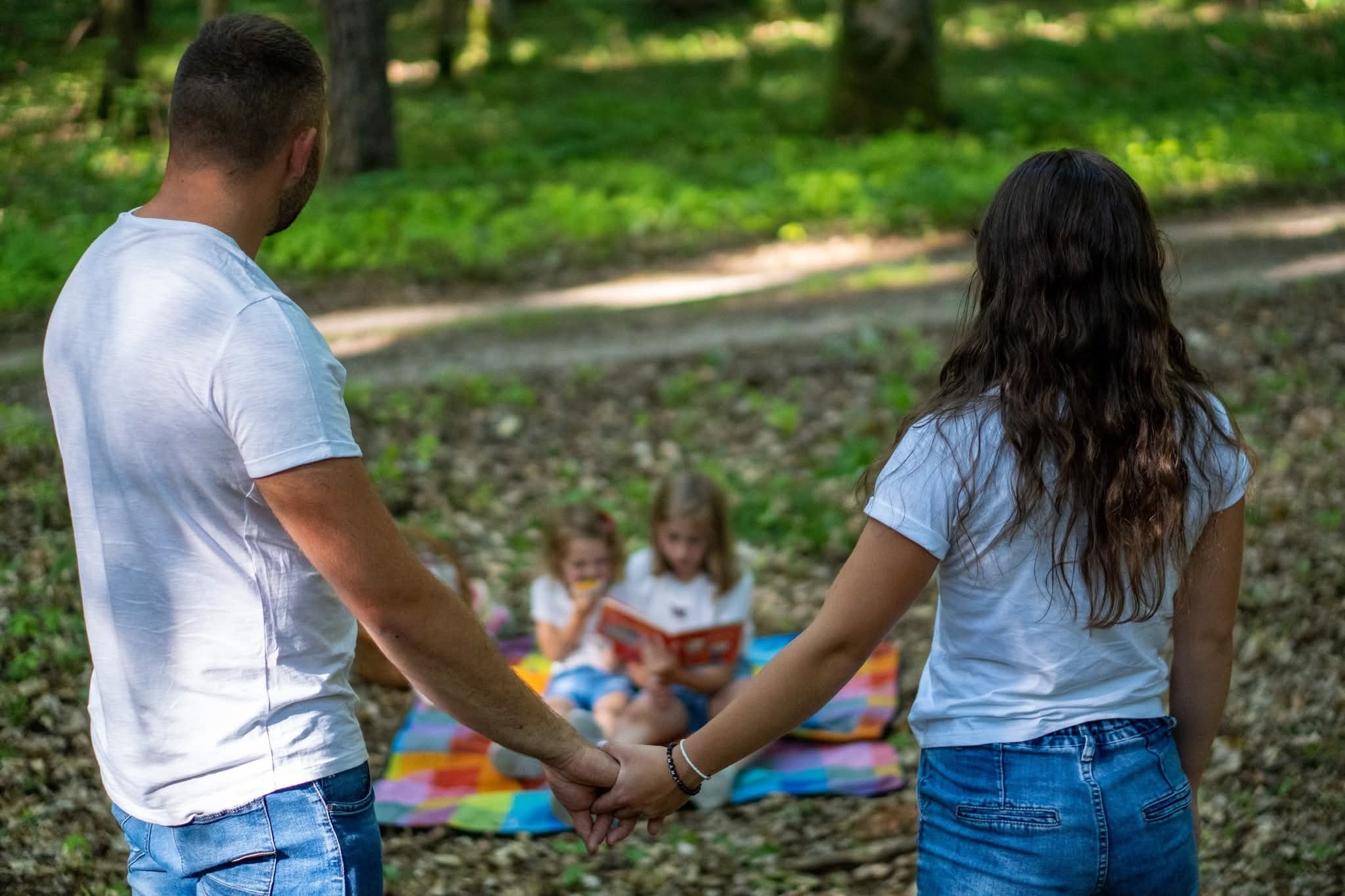 Para trzymająca się za ręce, z tyłu dwie dziewczynki czytają książkę na kolorowym kocu w lesie. Ujęcie zza pleców, naturalne światło.