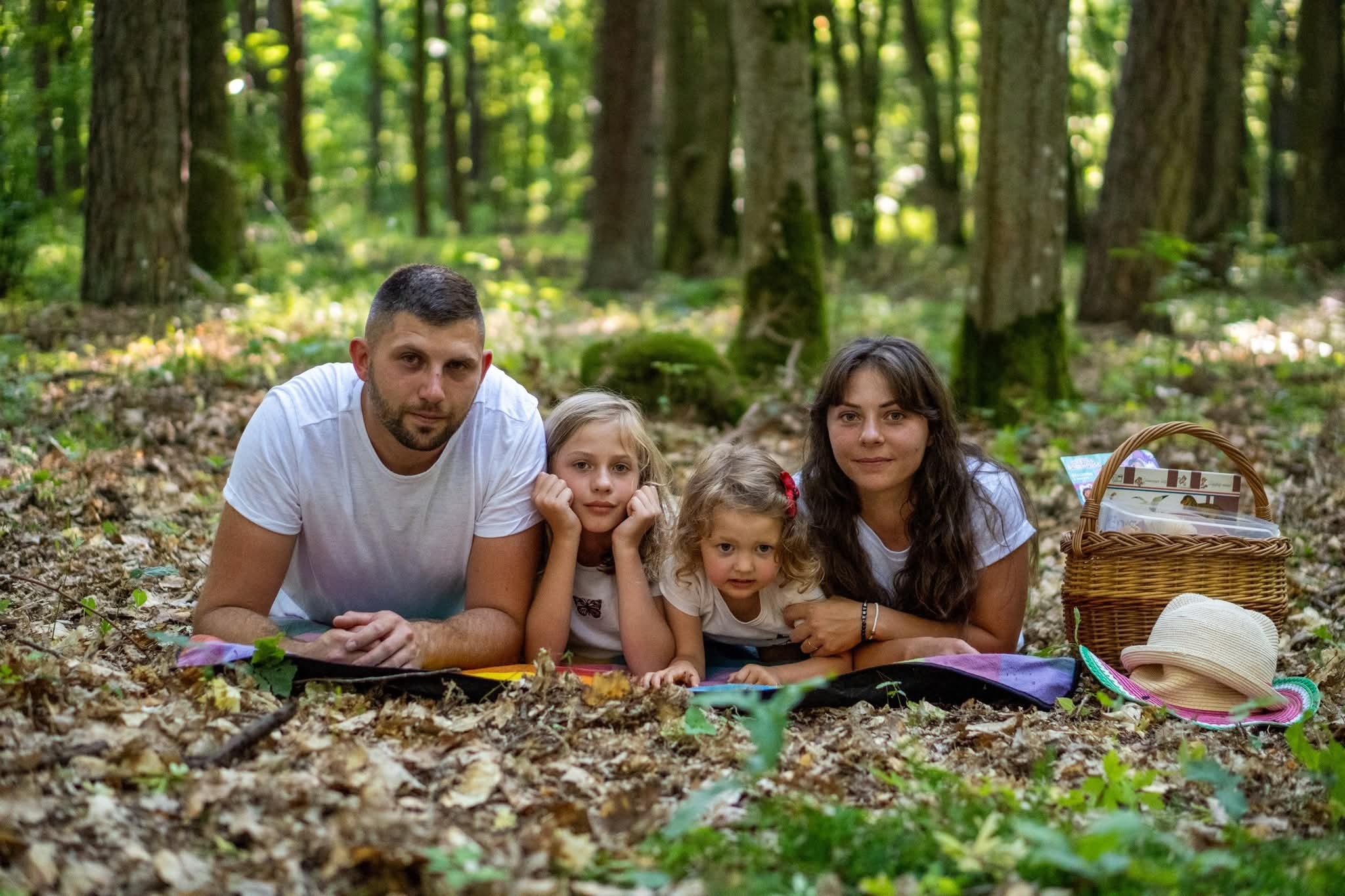 Rodzinne zdjęcie w lesie: tata, mama i dwie córki leżą na kocu piknikowym, obok koszyk piknikowy i kapelusz. Ujęcie z poziomu ziemi, drzewa w tle.