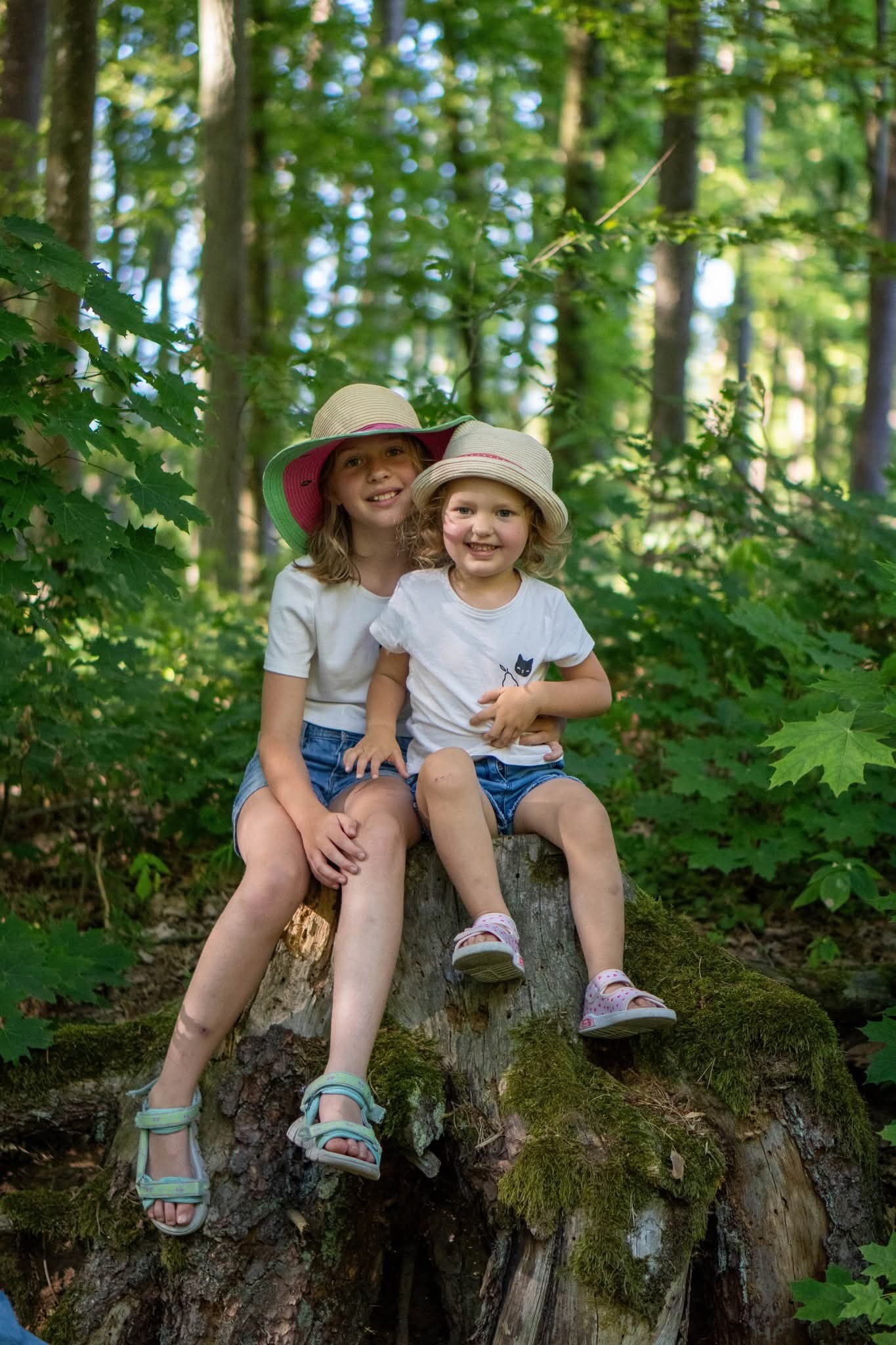 Dwie dziewczynki w kapeluszach siedzą na pniu drzewa w lesie. Dziewczynka w różowym kapeluszu obejmuje młodszą siostrę w białym kapeluszu. Naturalne światło, zielone tło.