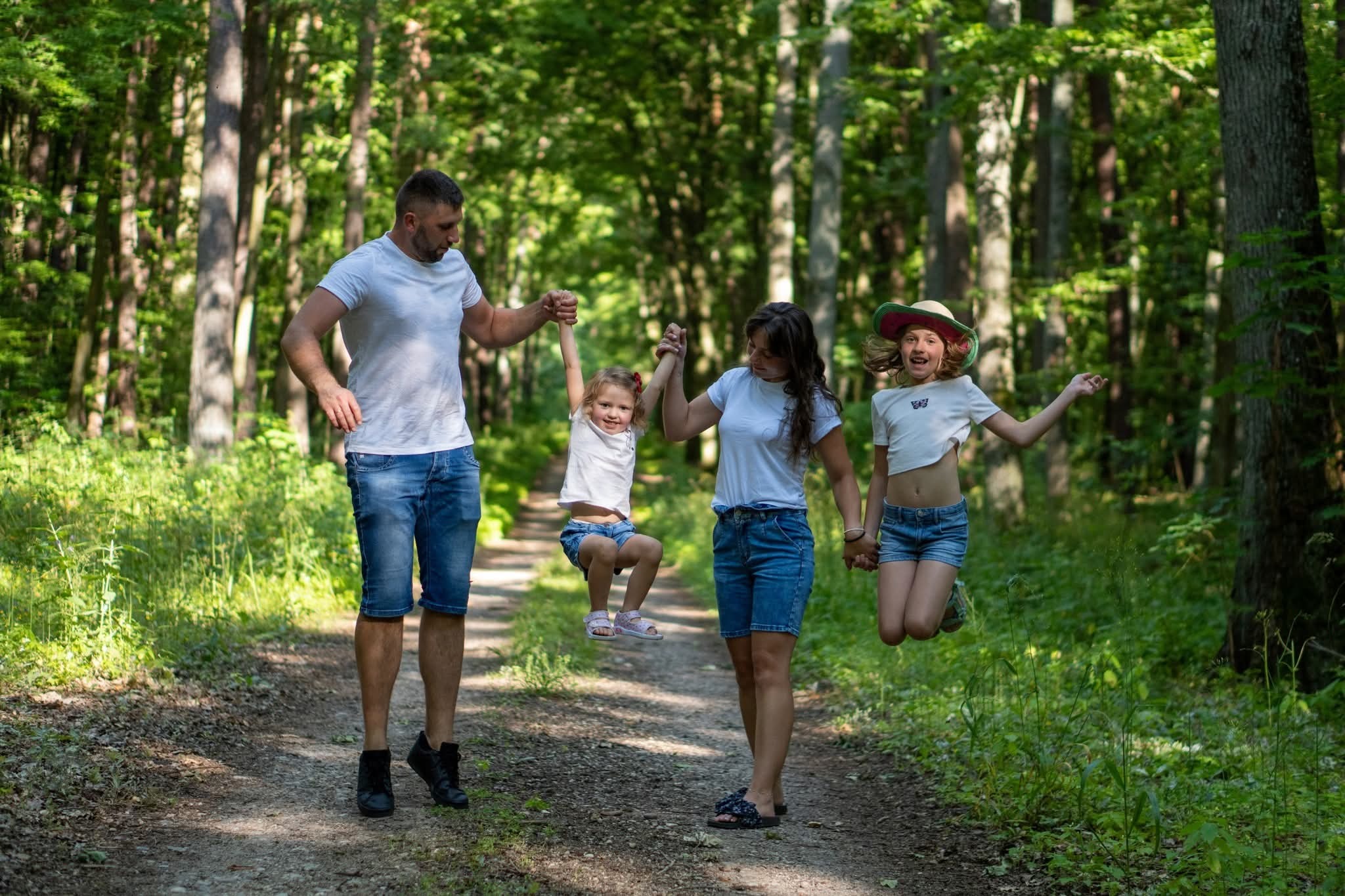 Uśmiechnięta rodzina na leśnej ścieżce: tata podnosi córkę, mama trzyma ją za rękę, a druga córka skacze. Wszyscy w letnich ubraniach, na tle zieleni drzew.