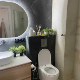 Nowoczesna łazienka z podświetlanym lustrem, umywalką na szafce z drewna, toaletą podwieszaną i szarą okładziną imitującą kamień na ścianie.