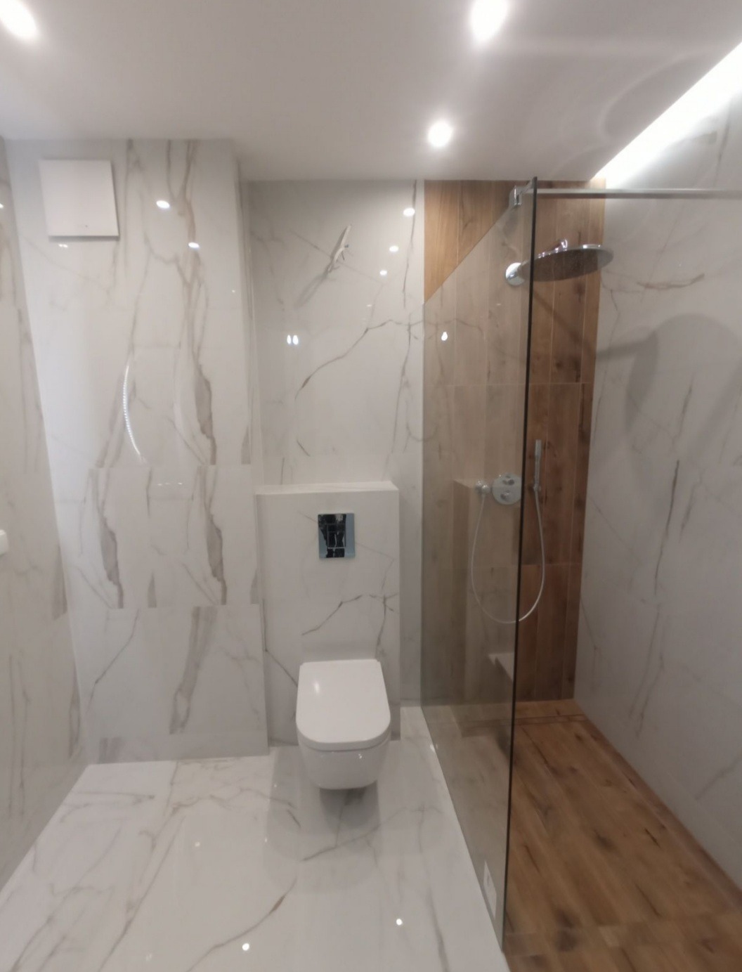 Nowoczesna łazienka z marmurowymi płytkami, podwieszaną toaletą i kabiną prysznicową typu walk-in z drewnianym akcentem. Minimalistyczny design, jasne oświetlenie.