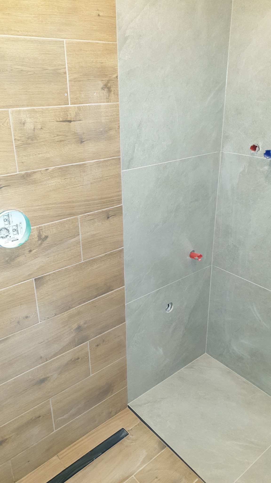 Wykończony fragment kabiny prysznicowej z dwoma rodzajami płytek: drewnopodobne poziome i szare, imitujące kamień. Widoczny odpływ liniowy i instalacja wodna.