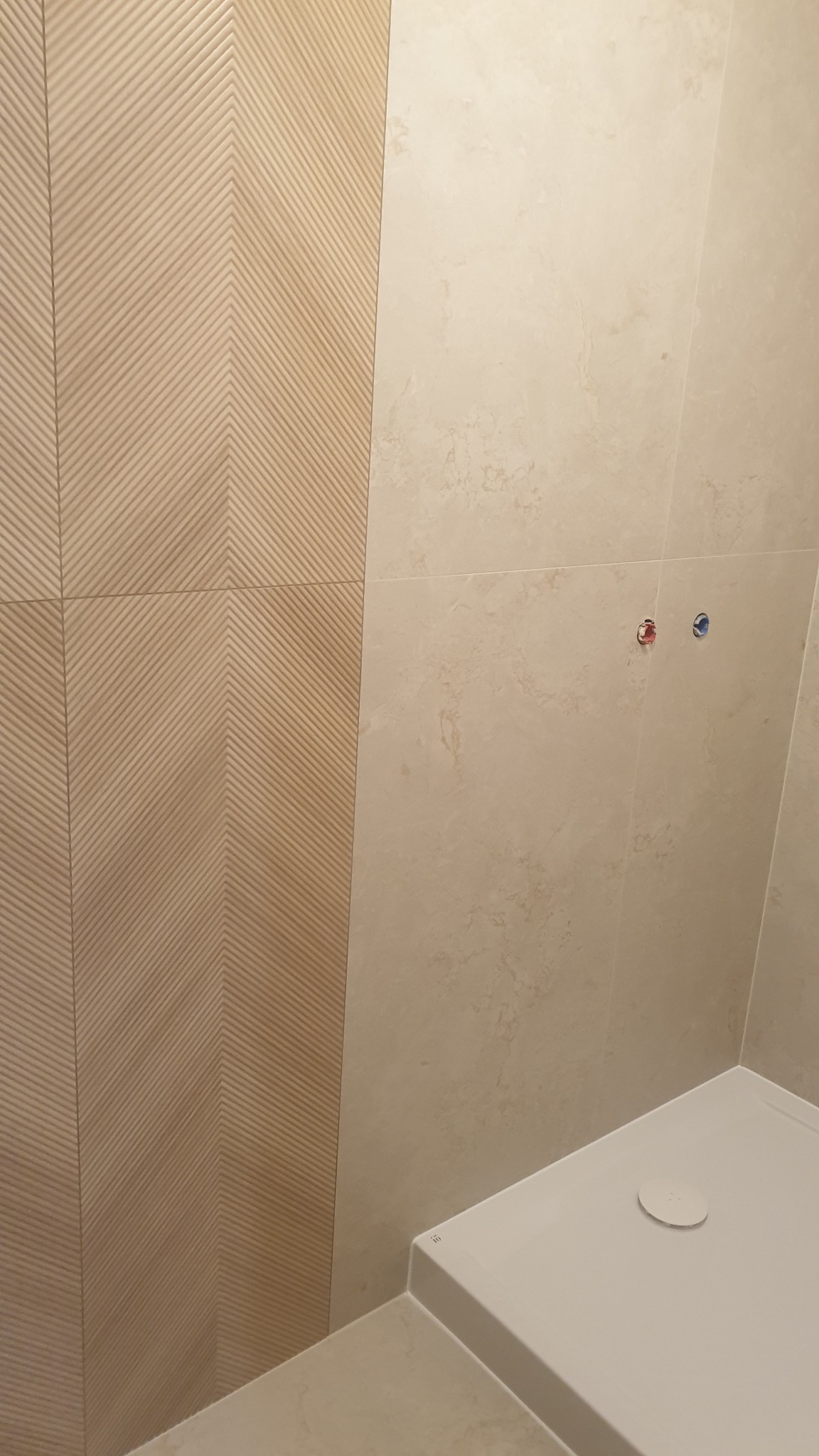 Beżowe płytki w łazience: ściana z wzorem chevron i gładkie, marmurkowe płytki. Widoczny brodzik i instalacja wodna.