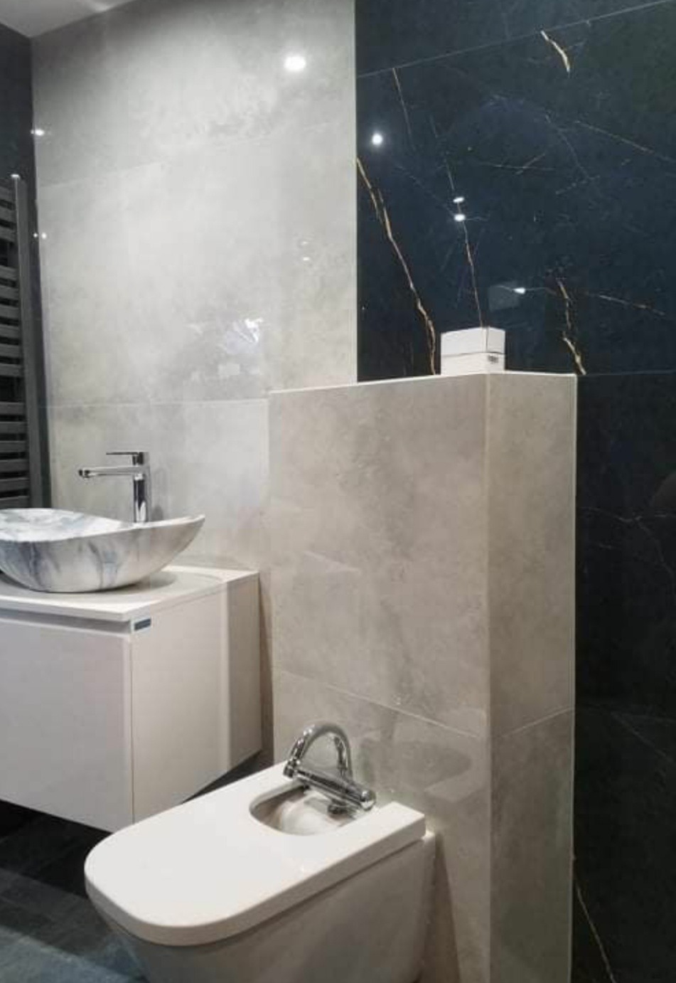 Nowoczesna łazienka z marmurową umywalką i czarnymi płytkami z złotymi żyłami, chromowana armatura, jasne płytki na ścianach i bidet wkomponowany w zabudowę.