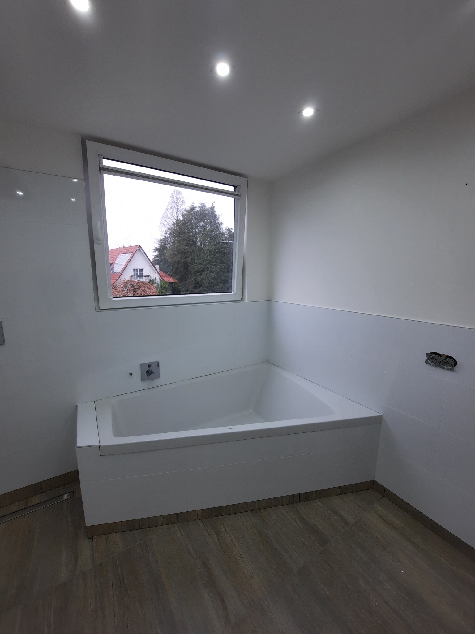 Biała wanna w wykończonej łazience z oknem, widok na dom i drzewa. Drewnopodobna podłoga, białe płytki do połowy ściany. Minimalistyczny design.