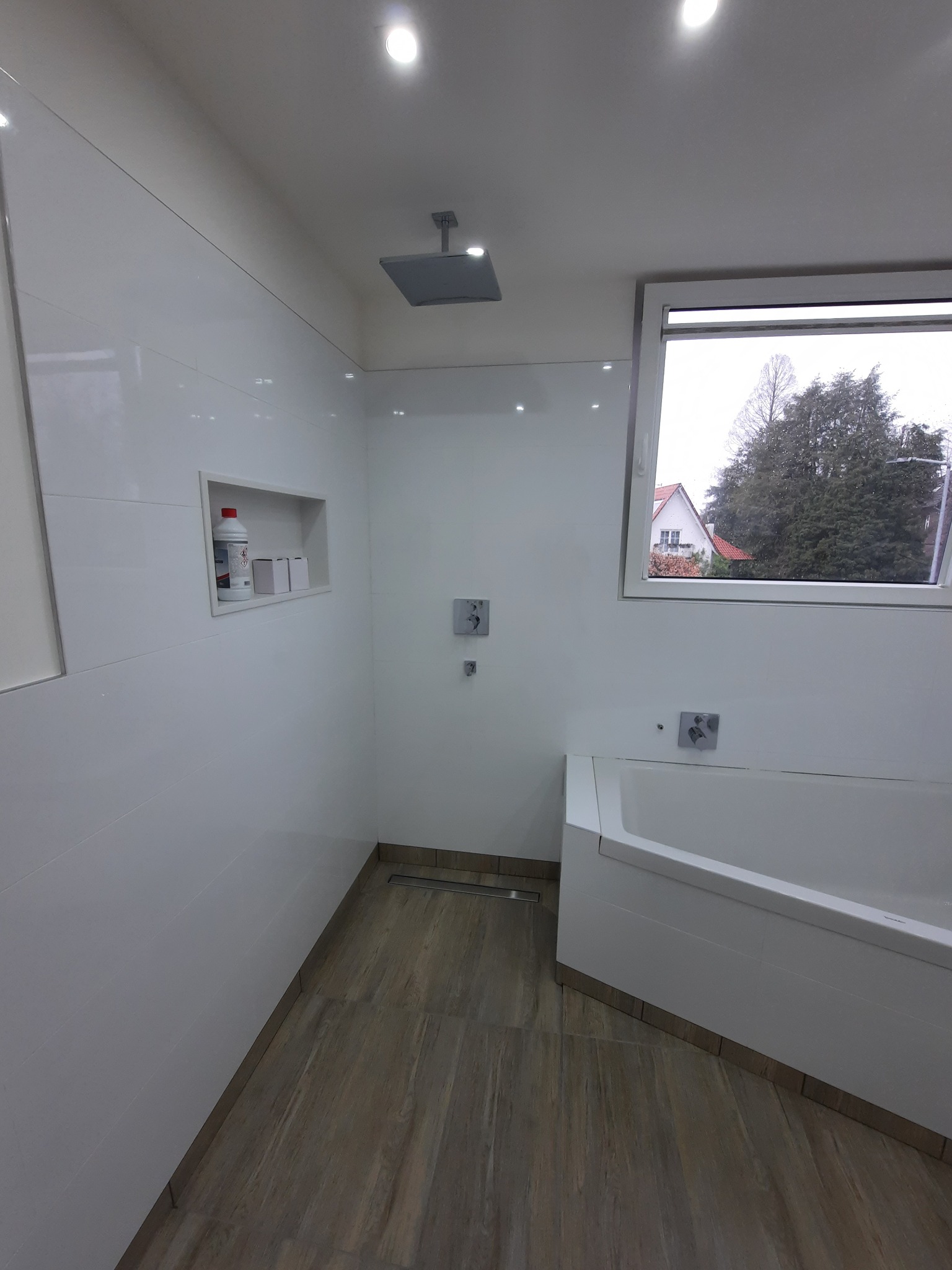 Biała łazienka po remoncie: płytki na ścianach i podłodze imitujące drewno, kabina prysznicowa typu walk-in, okno z widokiem na zewnątrz. Wnęka z kosmetykami.