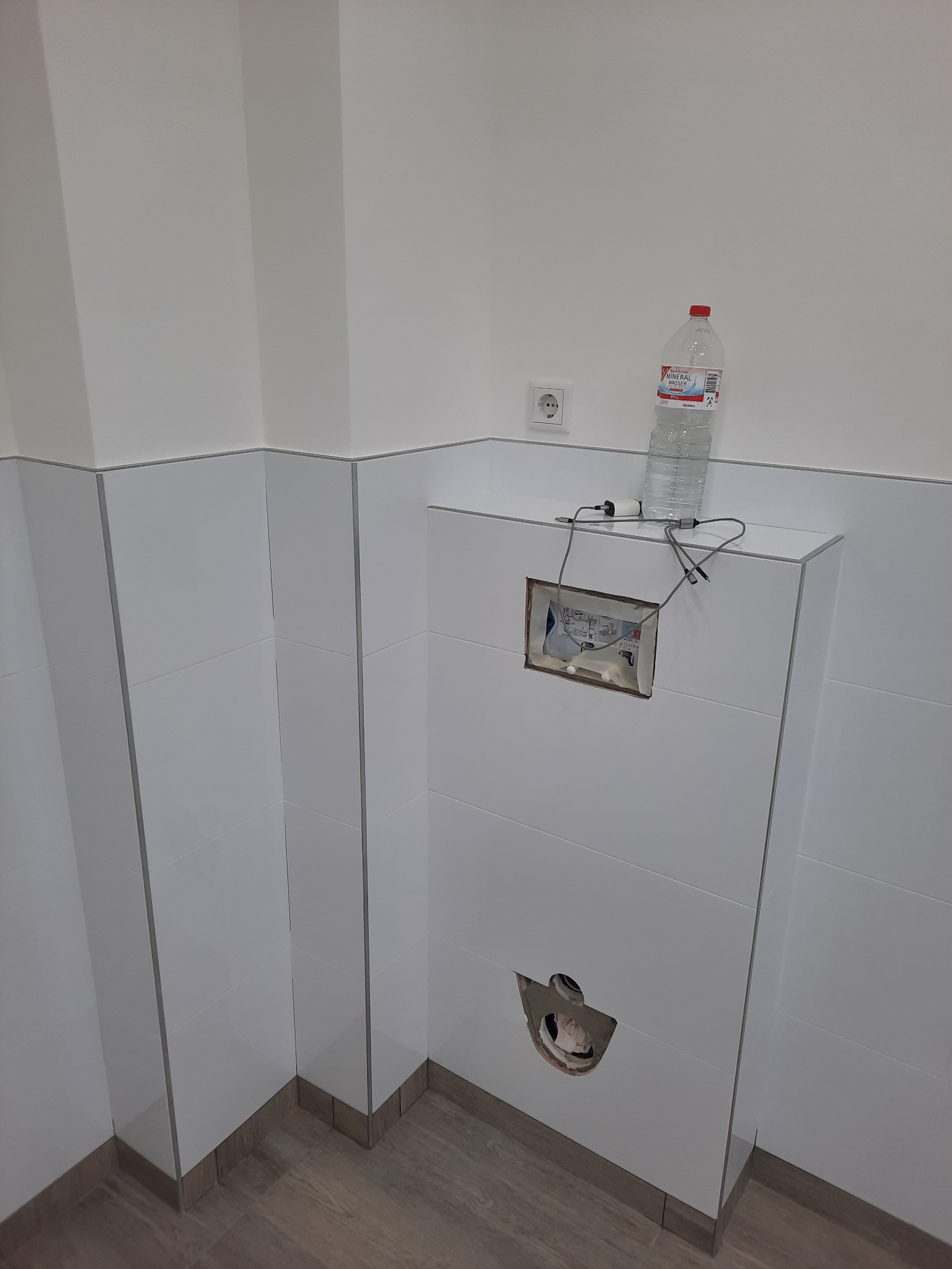 Biała zabudowa WC w trakcie montażu, widoczne otwory na przyciski i rury, butelka wody na górze. Minimalistyczna łazienka z drewnopodobną podłogą.