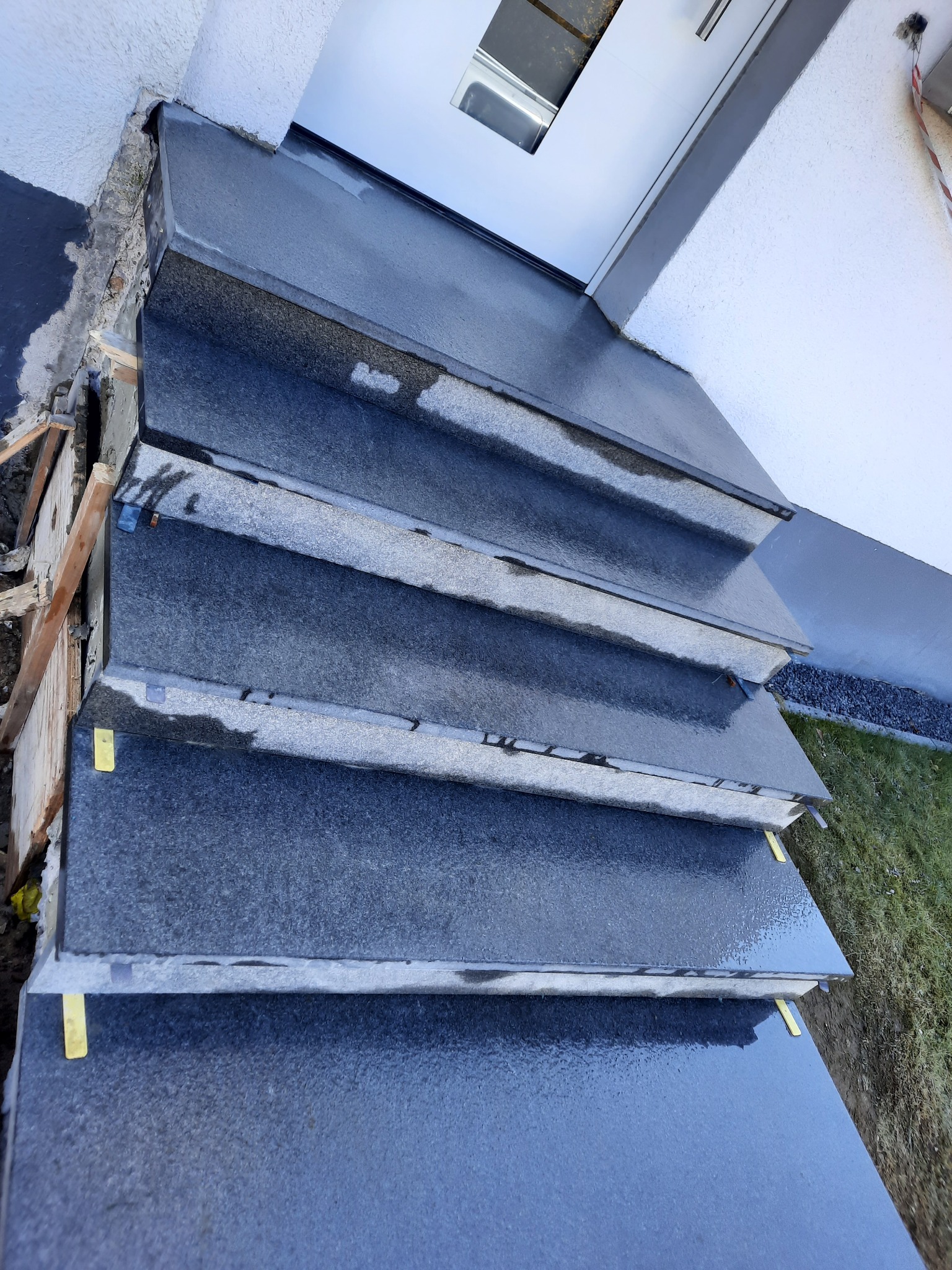 Granitowe schody zewnętrzne, świeżo po montażu, z widocznymi elementami mocującymi i fragmentem elewacji budynku. Nowoczesny wygląd, wysoki połysk kamienia.