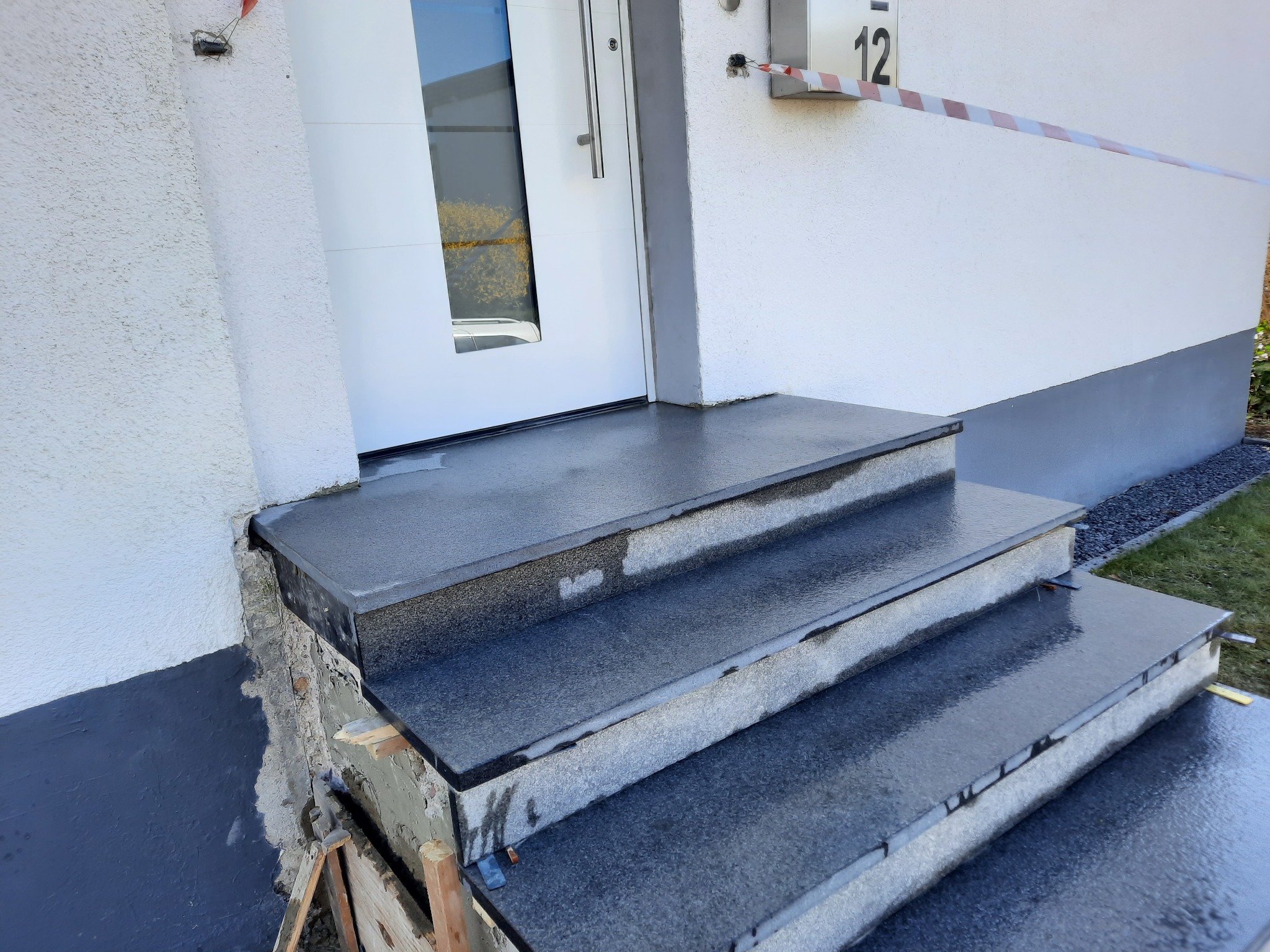 Granitowe schody wejściowe do domu w trakcie montażu, widoczne deski szalunkowe i fragment elewacji z numerem 12. Nowoczesny design.