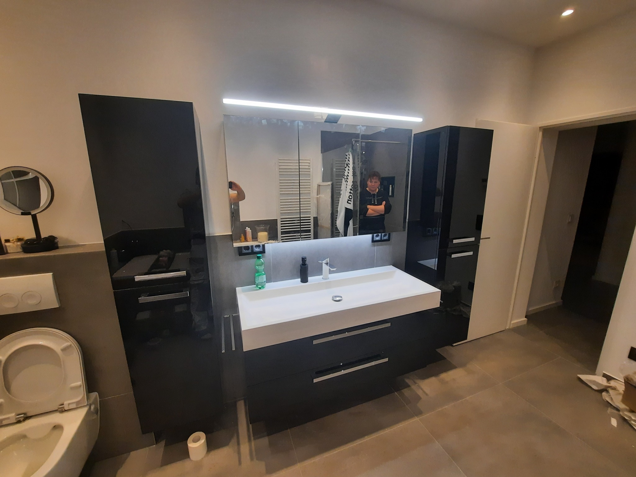 Nowoczesna łazienka z czarnymi szafkami i białym blatem umywalki podświetlonym LED, lustro z odbiciem osoby, płytki podłogowe w odcieniach szarości.