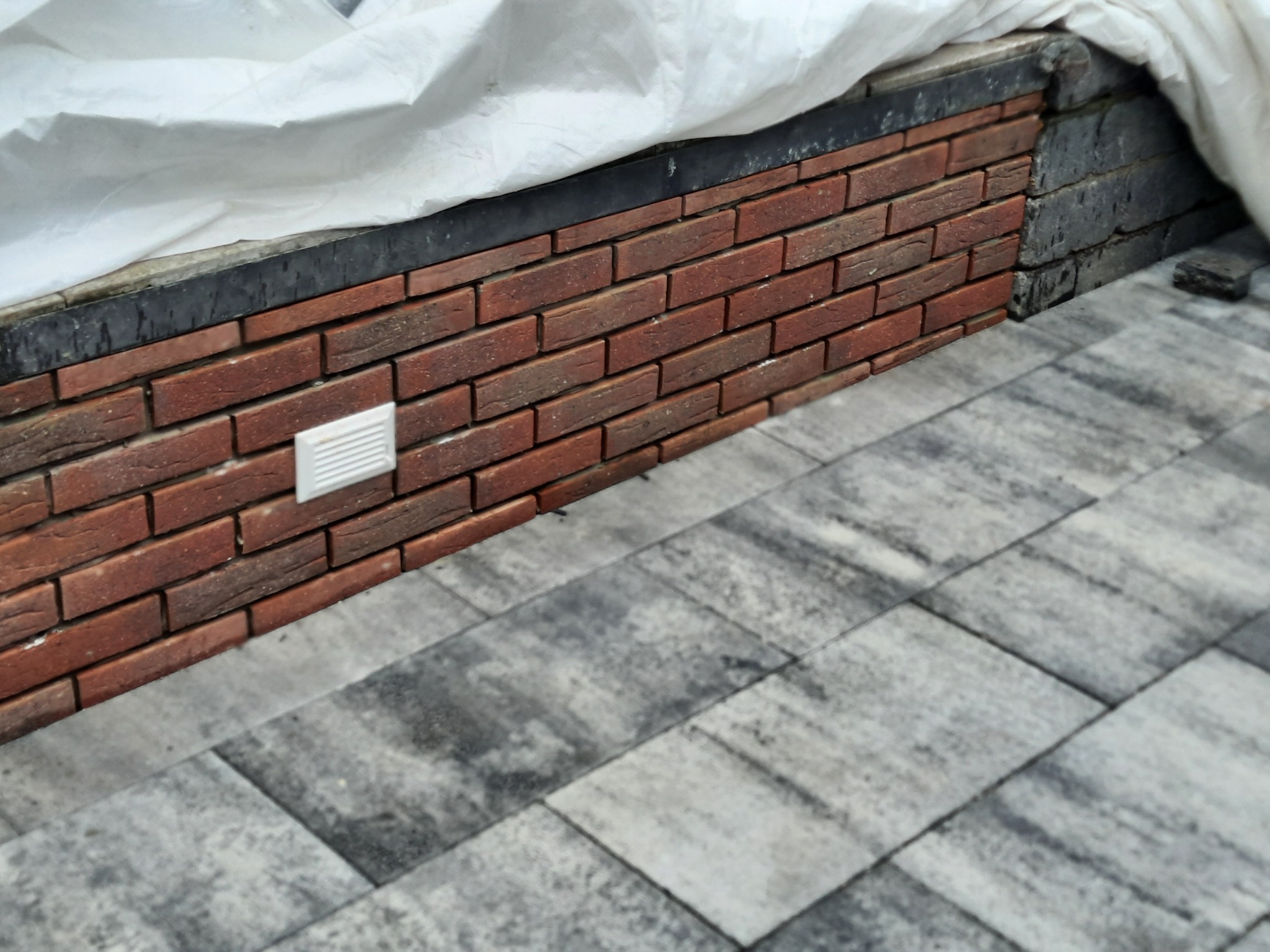 Murowana ściana z cegły klinkierowej z elementem elektrycznym, częściowo zakryta folią ochronną, sąsiadująca z szarą kostką brukową.