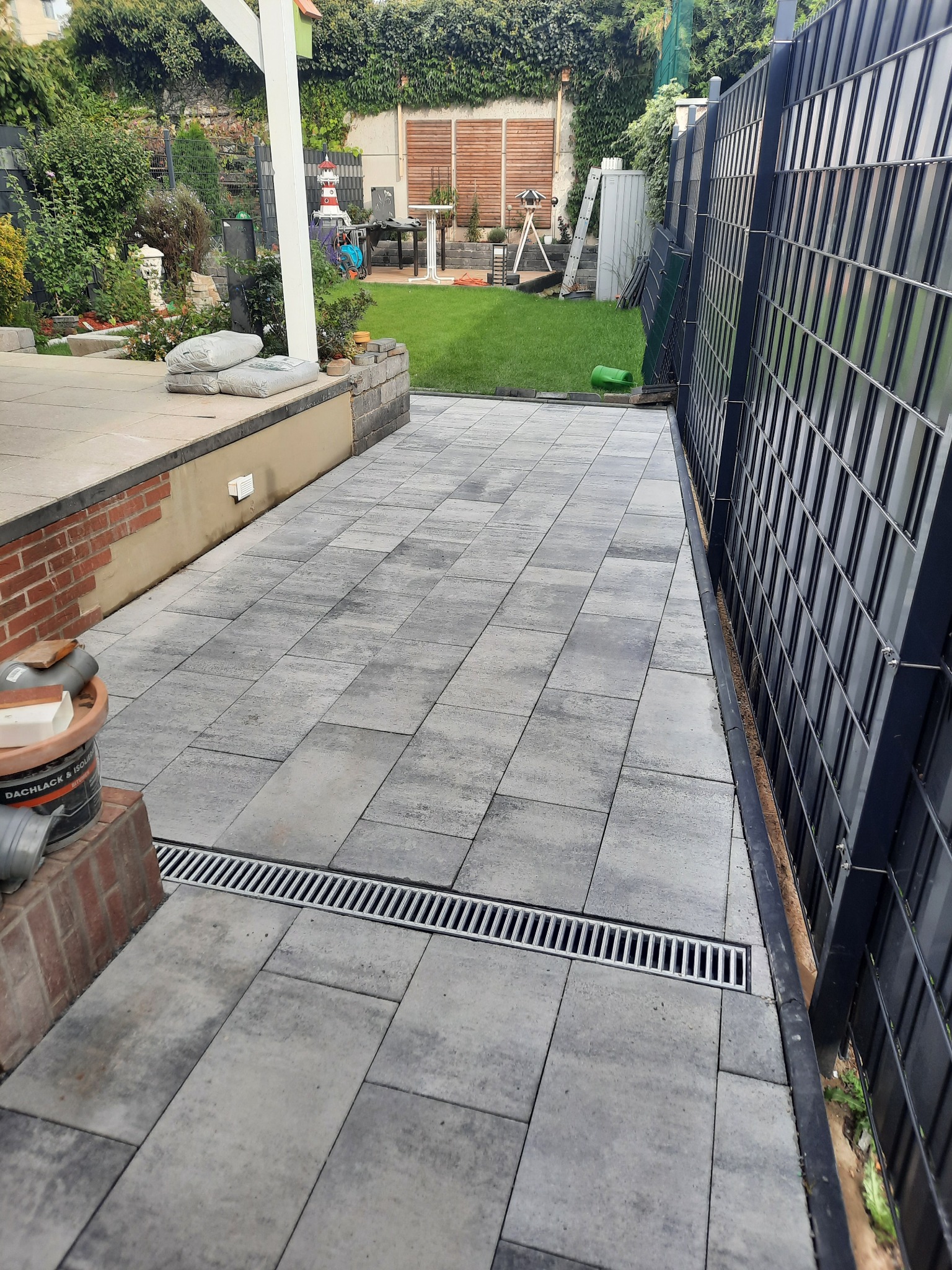 Ułożona szara kostka brukowa z odpływem liniowym na posesji, widoczne ogrodzenie panelowe i elementy ogrodu w tle.