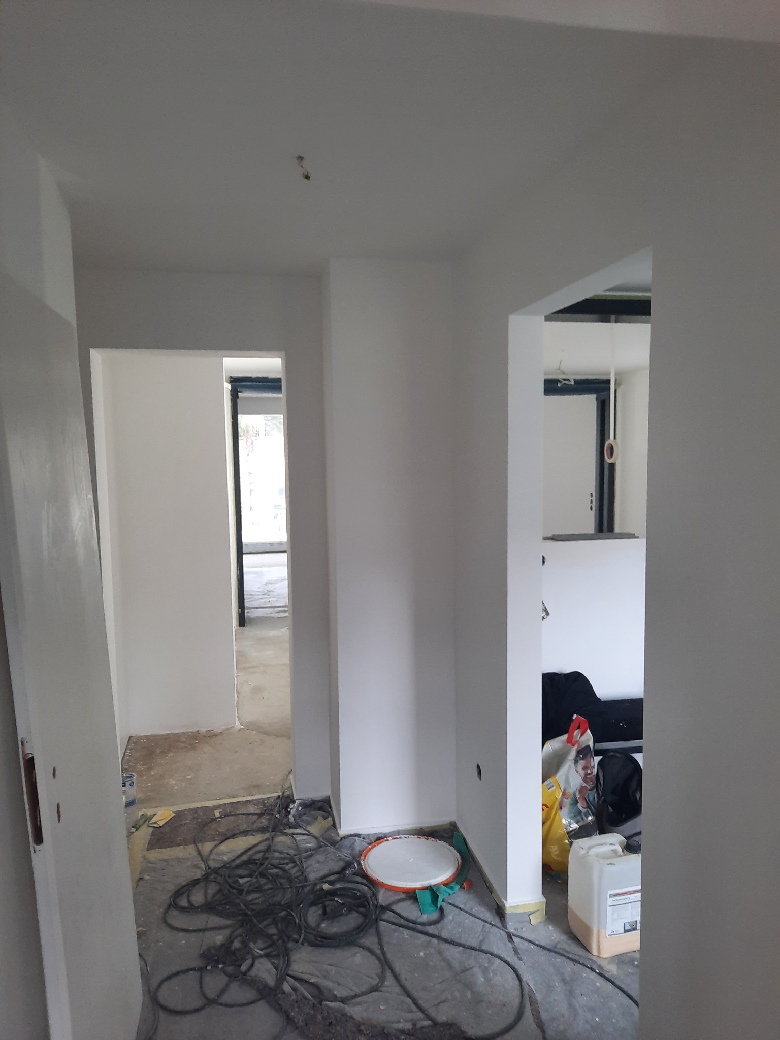 Wnętrze w trakcie malowania: świeżo pomalowane ściany na biało, widoczne kable, wiadro i akcesoria malarskie na podłodze zabezpieczonej folią malarską.