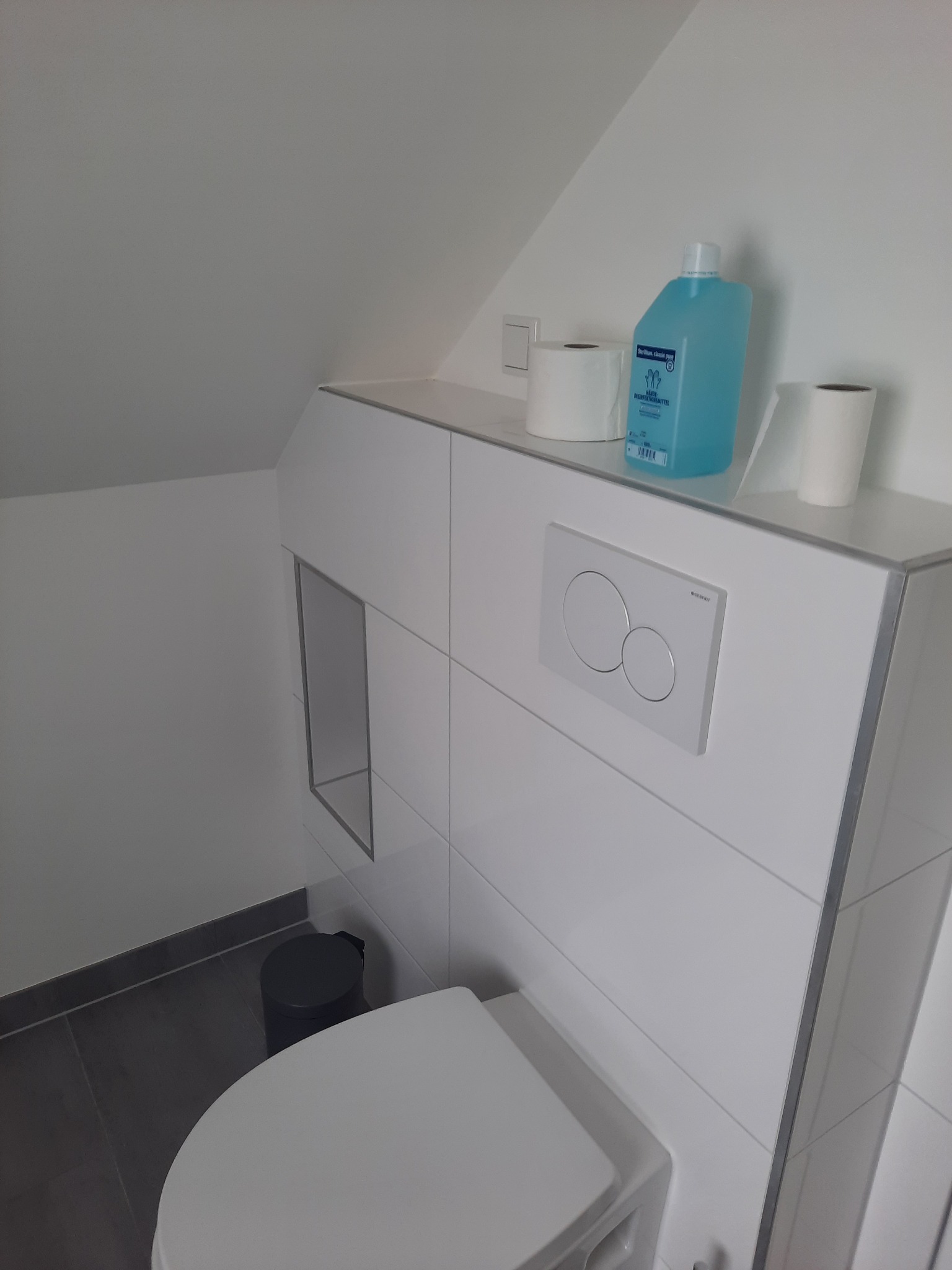 Nowoczesna toaleta podwieszana w łazience ze skośnym sufitem, z białymi płytkami, ukrytym zbiornikiem i niebieskim płynem dezynfekującym na blacie.