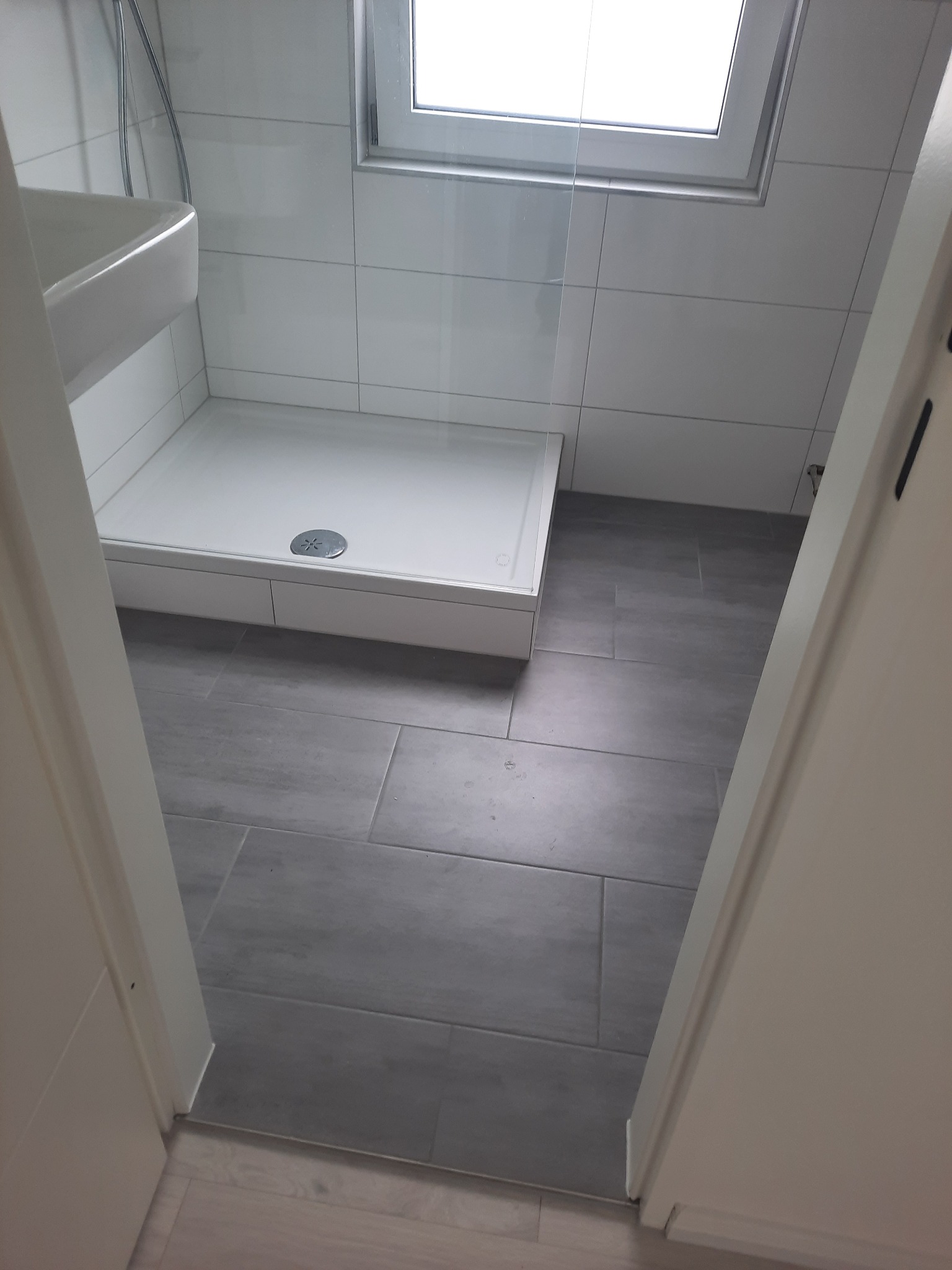 Nowoczesna łazienka z białą kabiną prysznicową i szarymi płytkami podłogowymi, widok zza uchylonych białych drzwi. Minimalistyczny design z oknem dachowym.