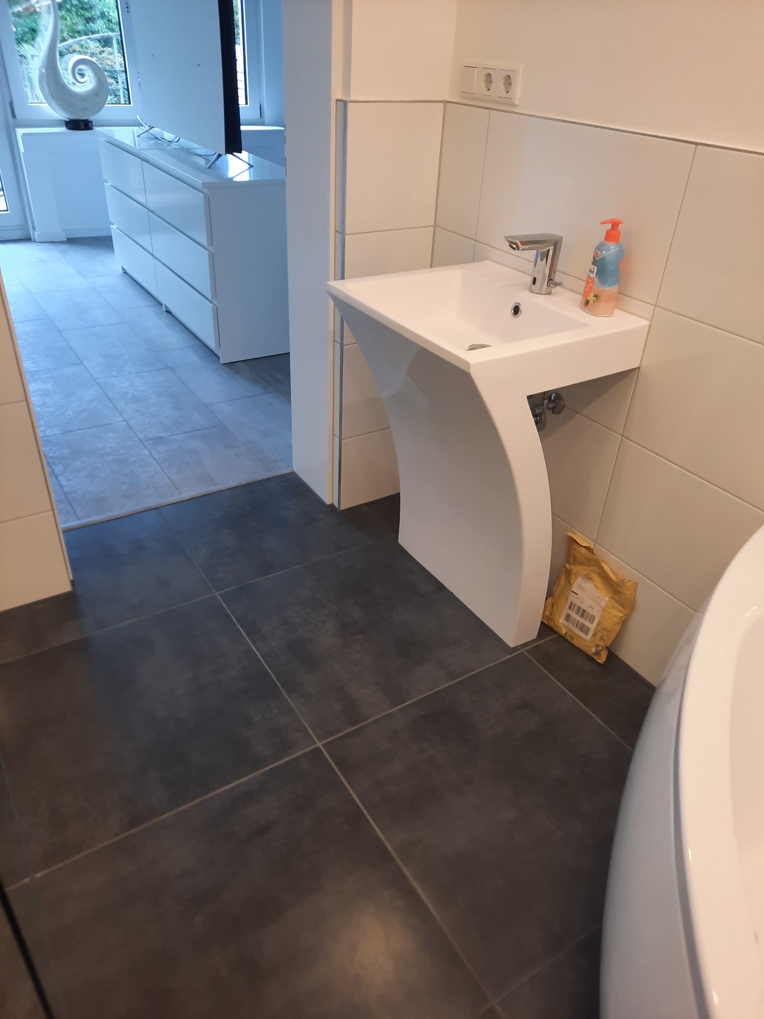 Nowoczesna umywalka z białą podstawą o zaokrąglonym kształcie w łazience z ciemnymi płytkami. W tle widoczny fragment wanny i przejście do innego pomieszczenia.