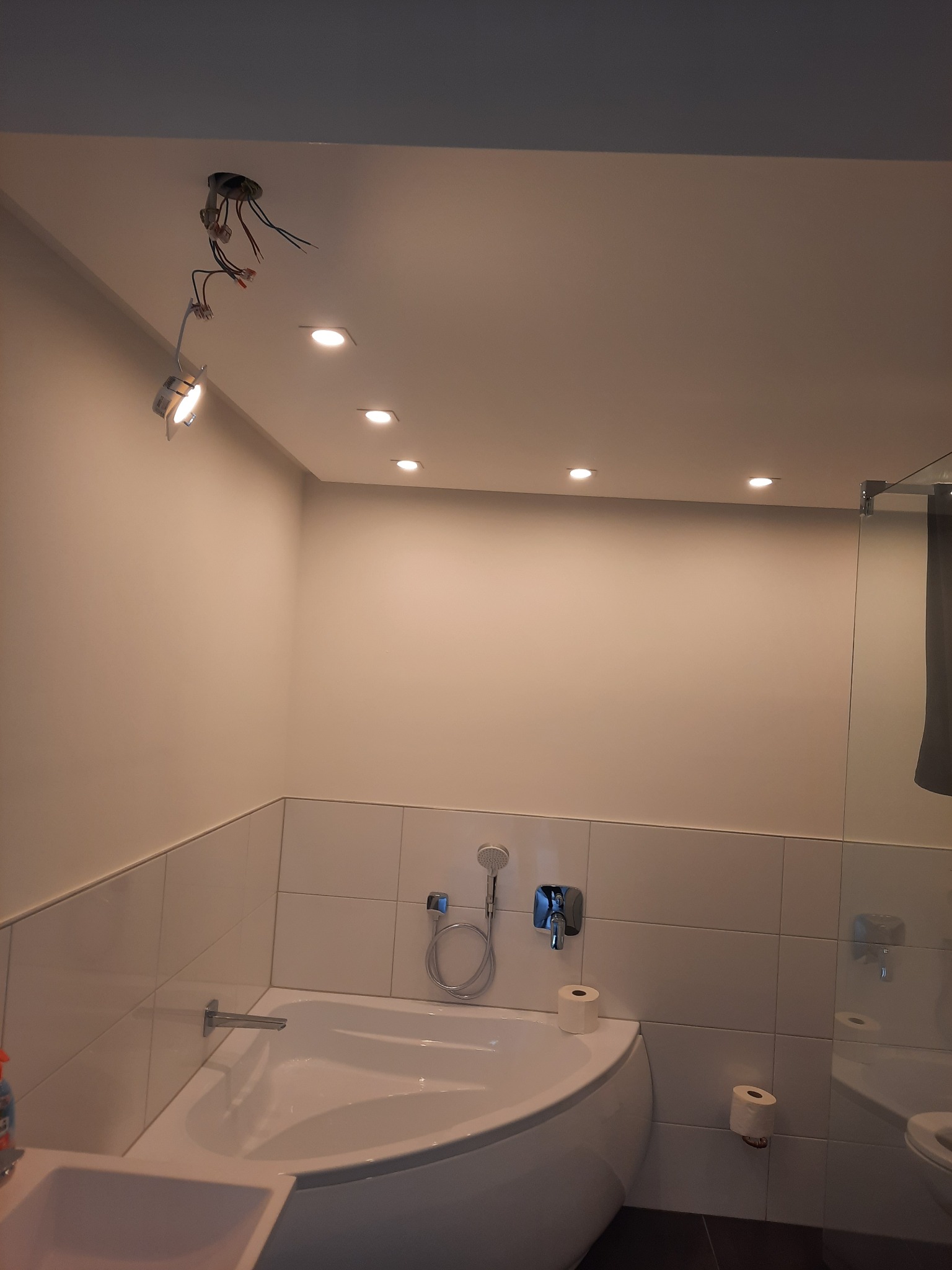 Nowoczesna łazienka w trakcie montażu oświetlenia: widoczne przewody i punktowe lampy sufitowe. Minimalistyczna wanna narożna i białe płytki.