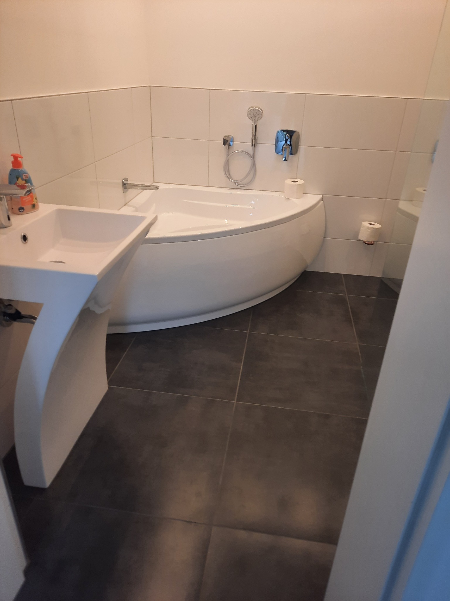 Biała, narożna wanna z chromowaną baterią i słuchawką prysznicową, obok biała umywalka na postumencie i szare płytki podłogowe w łazience.