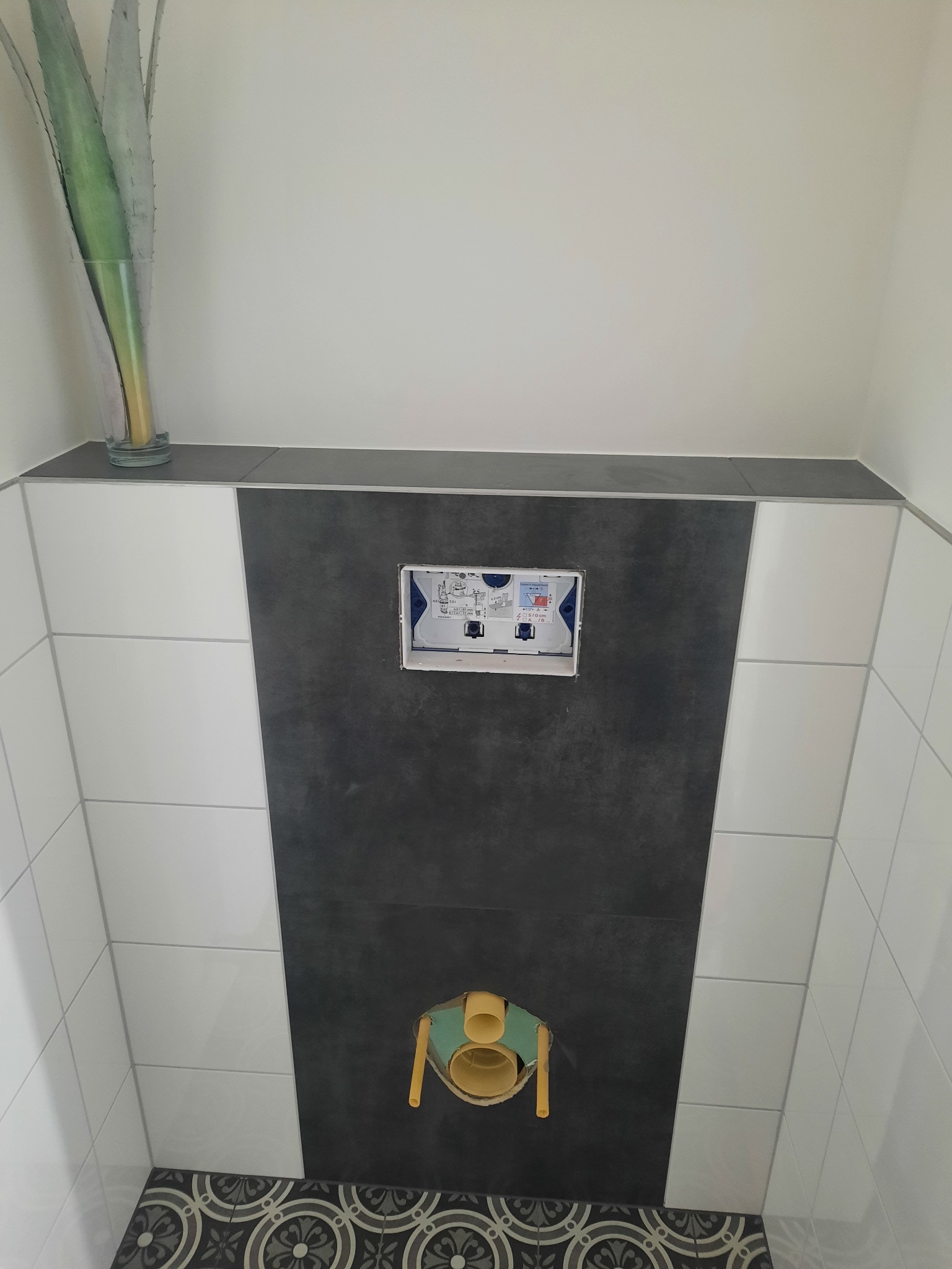 Wnęka z instalacją podtynkową WC na tle ciemnej płytki, obok białe płytki i dekoracyjna podłoga. W tle wazon z rośliną. Etap prac w łazience.