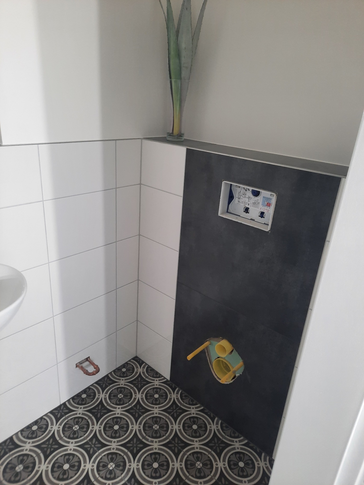 Fragment łazienki w trakcie wykańczania: białe i grafitowe płytki ścienne, dekoracyjna podłoga, widoczny stelaż podtynkowy WC i instalacja wodno-kanalizacyjna.