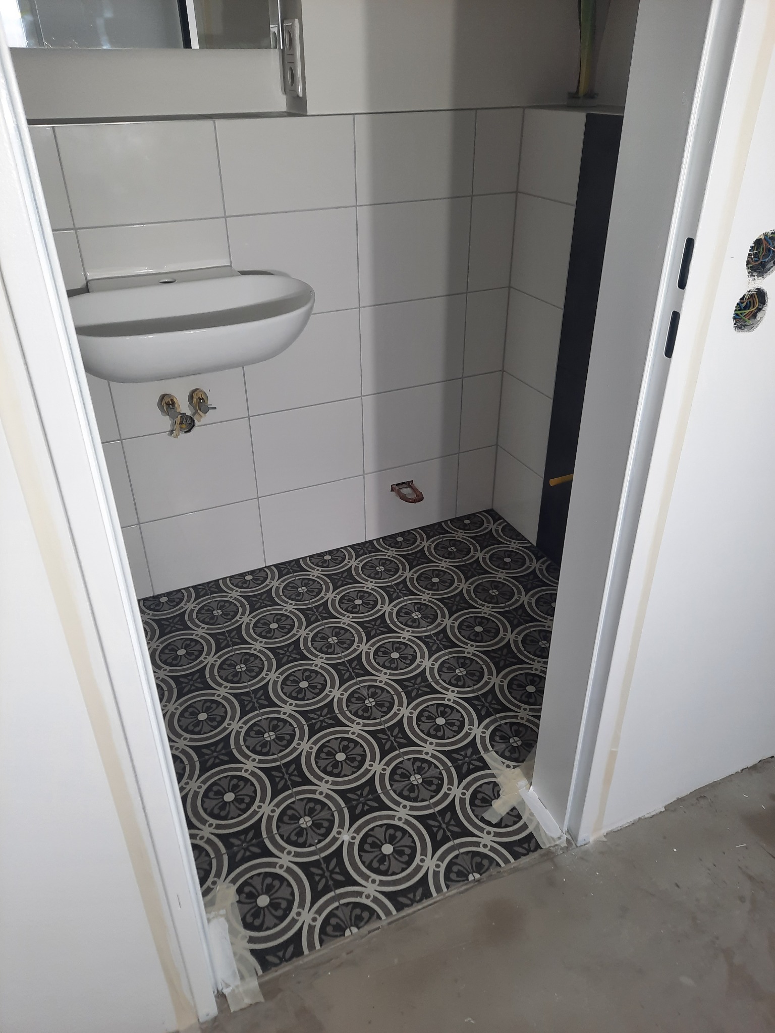 Biała umywalka na tle kwadratowych płytek, wzorzysta podłoga w łazience, widok przez otwarte drzwi. Wnętrze w trakcie wykończenia, widoczne elementy instalacji.