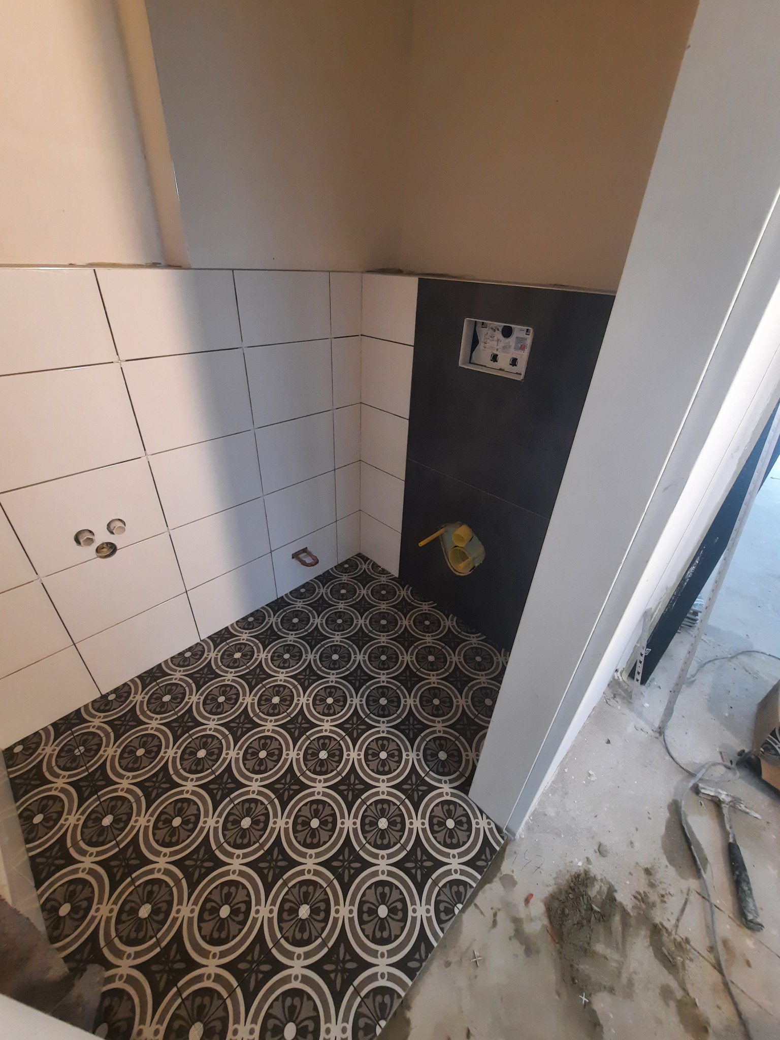 Częściowo wykończona łazienka z białymi i czarnymi płytkami ściennymi oraz wzorzystą podłogą. Widoczne narzędzia glazurnicze.