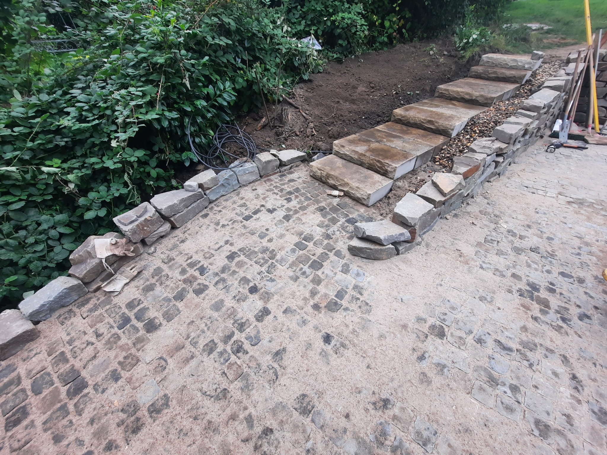 Ułożona kostka brukowa, kamienne schody tarasowe w ogrodzie, obrzeże z kamienia. Widoczne narzędzia budowlane.