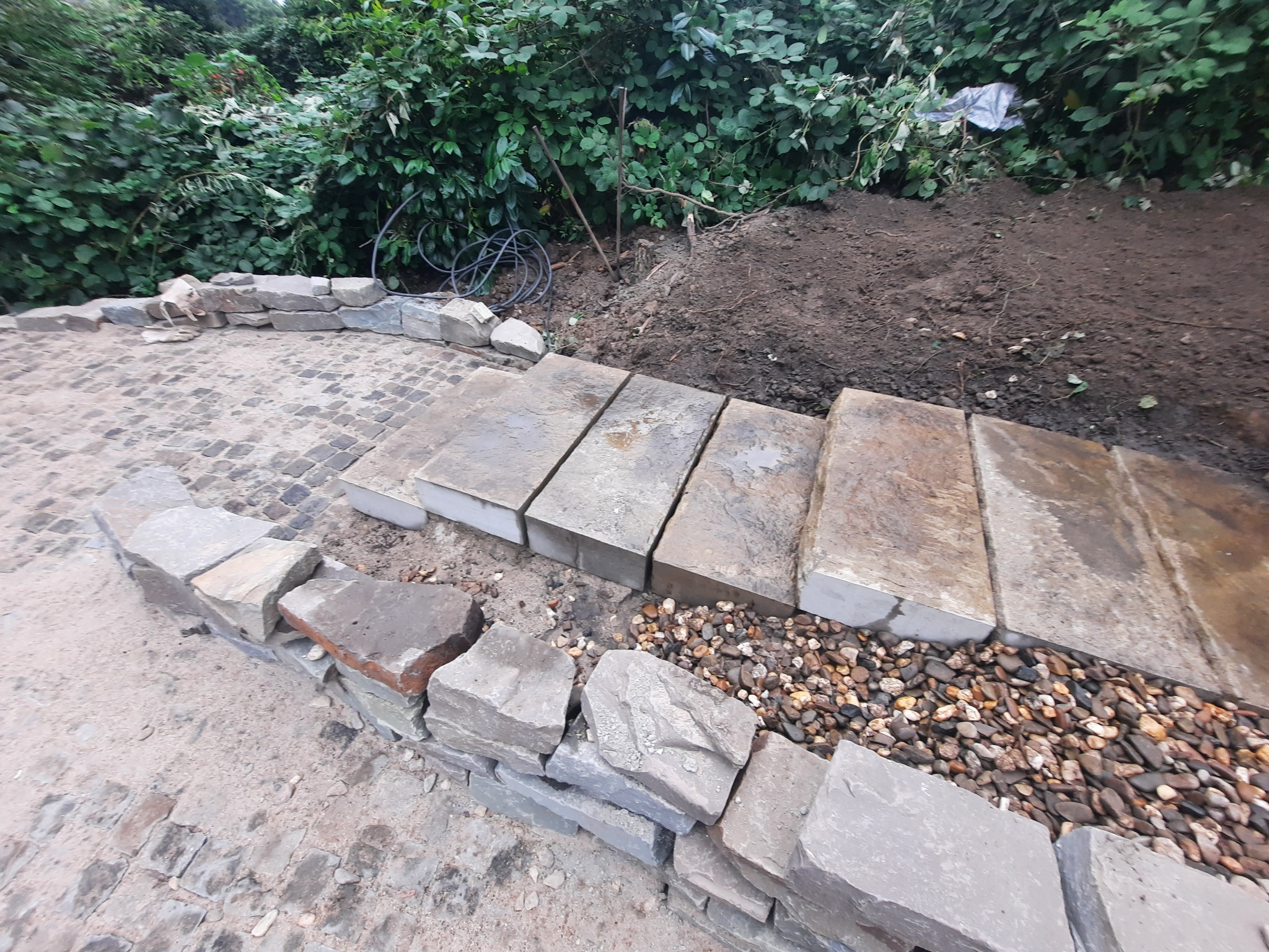 Kamienne schody ogrodowe w trakcie budowy, z widocznym kamiennym obrzeżem i przygotowanym podłożem, otoczone zielenią. Detal układania kamiennych elementów.