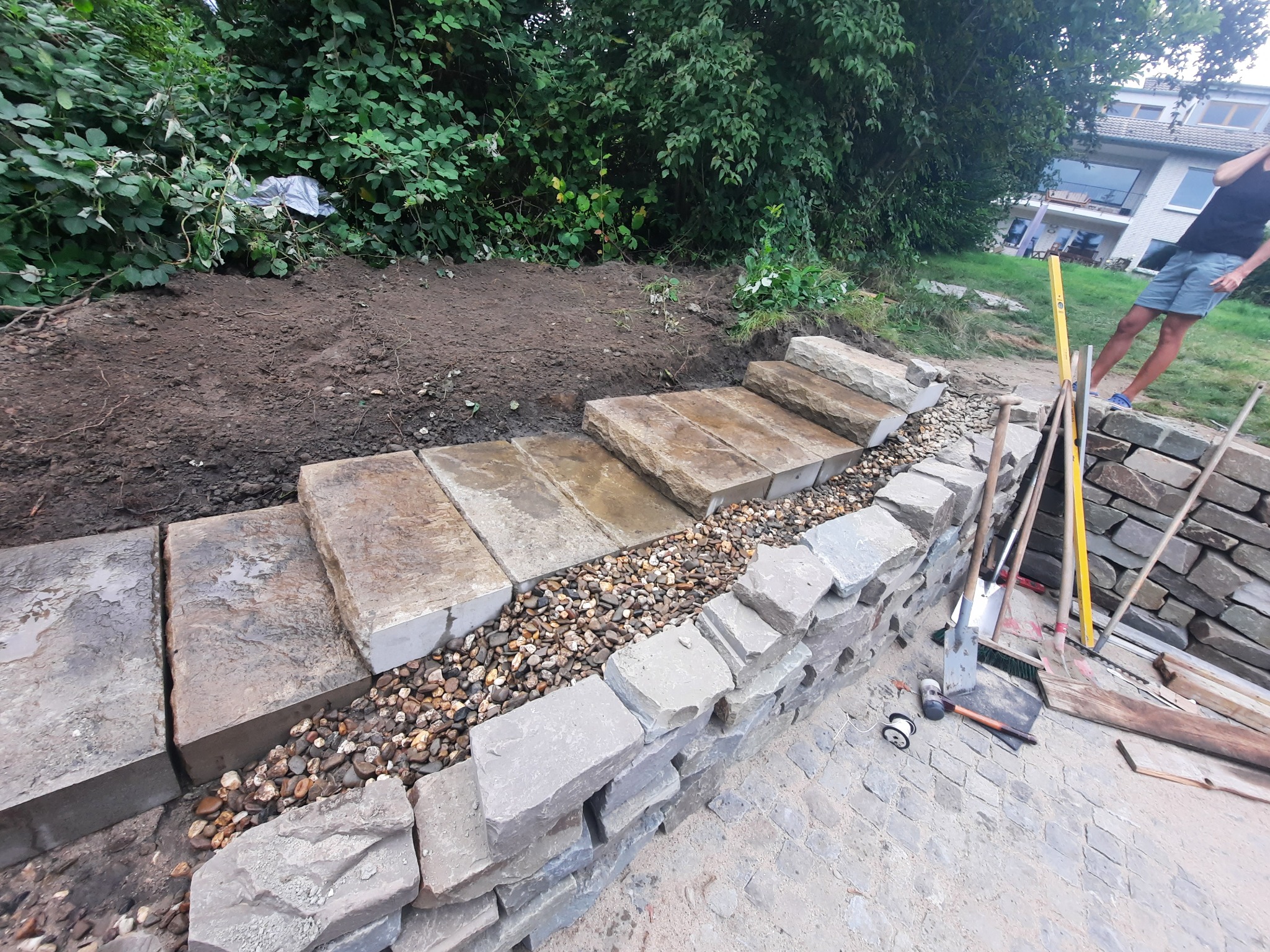 Budowa schodów zewnętrznych z kamienia: stopnie z kamiennych płyt, wypełnienie z drobnych kamyków, mur z nieregularnych kamieni. Widoczne narzędzia i osoba w tle.