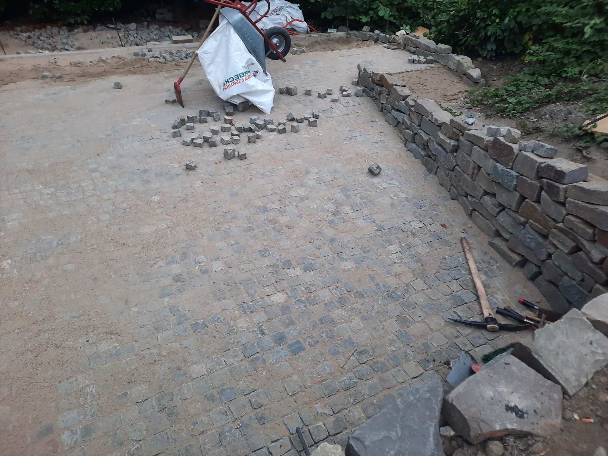 Układanie kostki brukowej na posesji: widoczna ułożona część nawierzchni, materiały budowlane, narzędzia (szpadel, kilof) i fragment murku z kamienia.