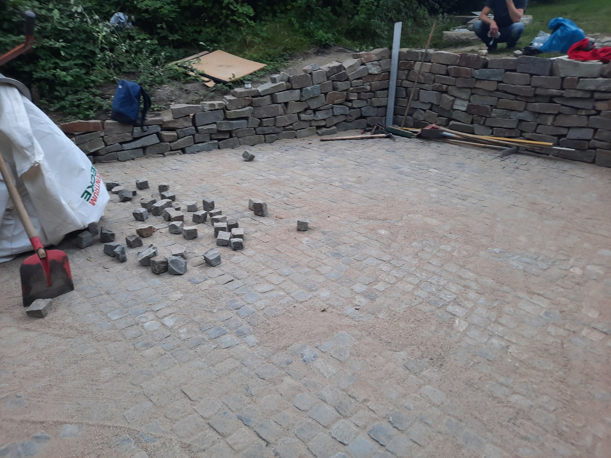 Układanie kostki brukowej na podsypce piaskowej, widoczna część murku z kamienia, narzędzia i materiały budowlane. Prace w toku, etap przygotowania podłoża.
