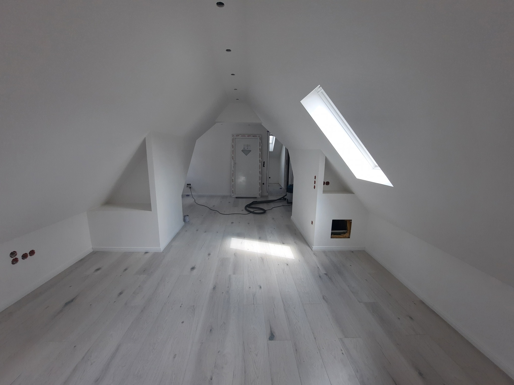 Jasne poddasze po remoncie: białe ściany, okno dachowe i panele podłogowe w odcieniach szarości. Widoczne punkty oświetleniowe w suficie i gniazdka elektryczne.