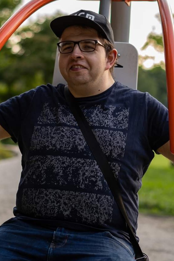 Mężczyzna w czapce i okularach ćwiczy na siłowni plenerowej, ubrany w ciemną koszulkę z wzorem i jeansy. Pomarańczowe elementy konstrukcji, zielone tło drzew.