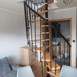 Interior Wykończenia Wnętrz Łukasz Chudzik - Spiralne schody z drewnianymi stopniami i czarną metalową konstrukcją w jasnym wnętrzu domu w Jastrzębiu. Widok na łazienkę w tle.