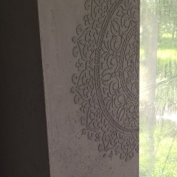 Interior Wykończenia Wnętrz Łukasz Chudzik - Dekoracyjna ściana z ornamentem w stylu mandali, wykonana z szarego tynku strukturalnego, częściowo zasłonięta folią ochronną przy oknie.