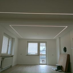 Interior Wykończenia Wnętrz Łukasz Chudzik - Nowoczesne oświetlenie LED w kształcie prostokątów na suficie w jasnym pokoju z dwoma oknami i parkietem. Karton oparty o ścianę.