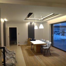 Interior Wykończenia Wnętrz Łukasz Chudzik - Jasne wnętrze jadalni z drewnianą podłogą, białym stołem i krzesłami w folii. Sufit podwieszany z oświetleniem LED. Duże okno z widokiem na zimowy krajobraz.