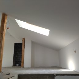 Interior Wykończenia Wnętrz Łukasz Chudzik - Wnętrze poddasza po malowaniu ścian na biało, z drewnianymi belkami i oknem dachowym. Widoczny surowy beton na podłodze i fragment instalacji elektrycznej.