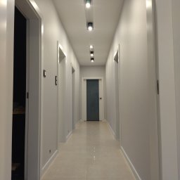 Interior Wykończenia Wnętrz Łukasz Chudzik - Długi, jasny korytarz z białymi drzwiami i punktowym oświetleniem sufitowym. Podłoga wyłożona jasnymi płytkami. Widok w głąb korytarza.
