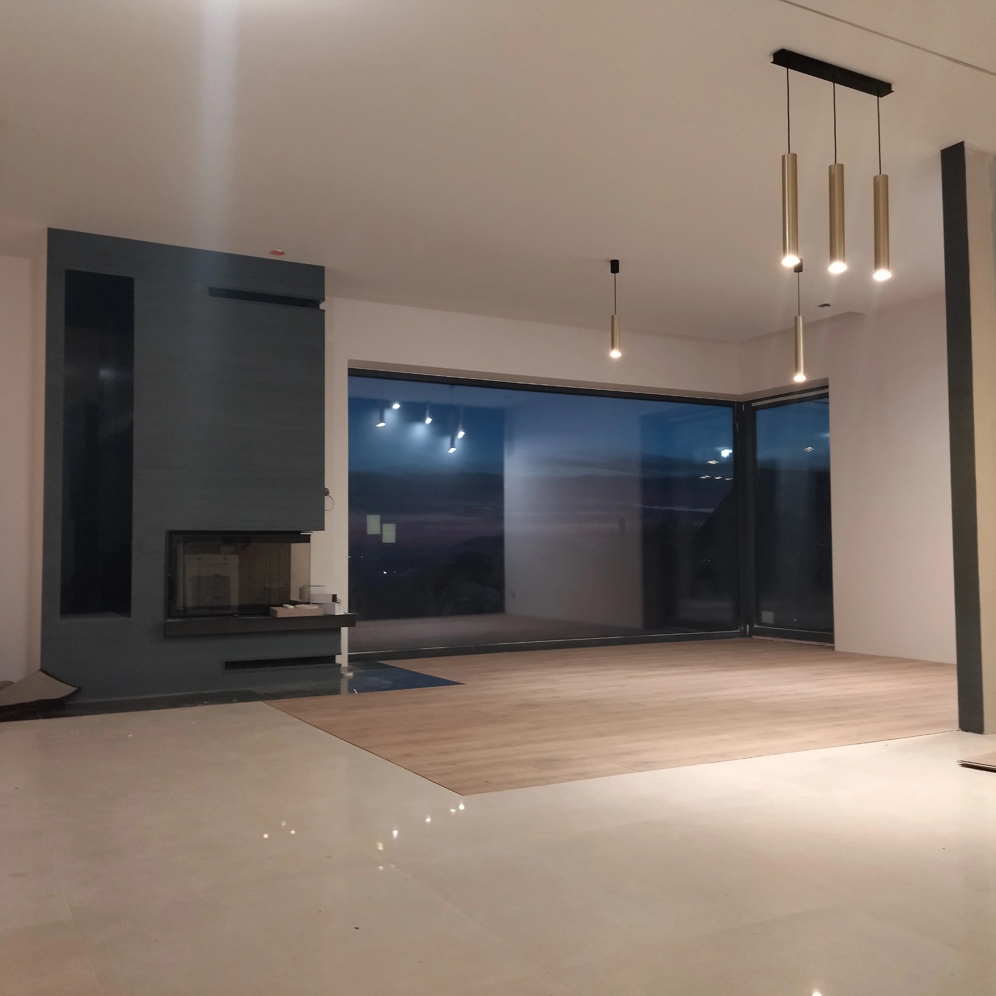 Salon z kominkiem i dużym oknem, częściowo wyłożony parkietem. Widoczne nowoczesne lampy sufitowe i minimalistyczny design wnętrza. Podłoga wyłożona jasnymi płytkami.