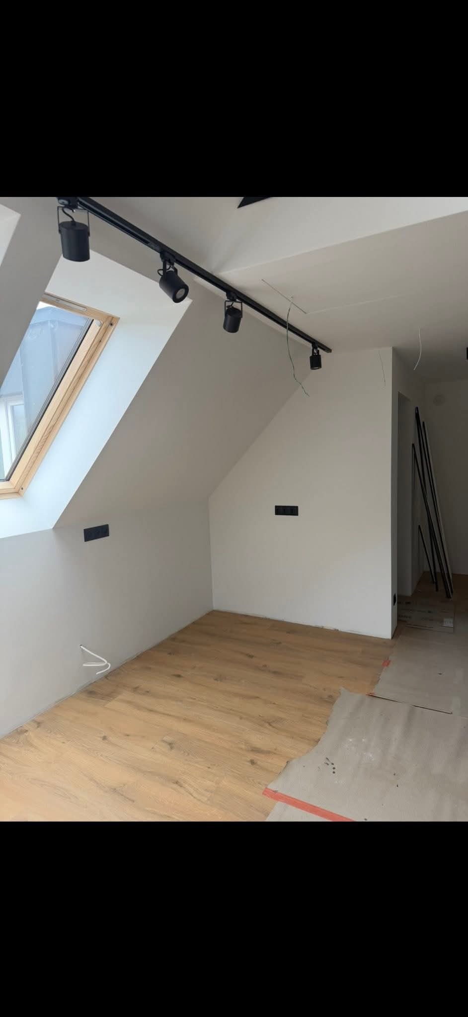 Poddasze z oknem dachowym i nową, jasną podłogą z paneli. Widoczne czarne reflektory na szynie i fragment przygotowanego podłoża pod dalszy montaż paneli.