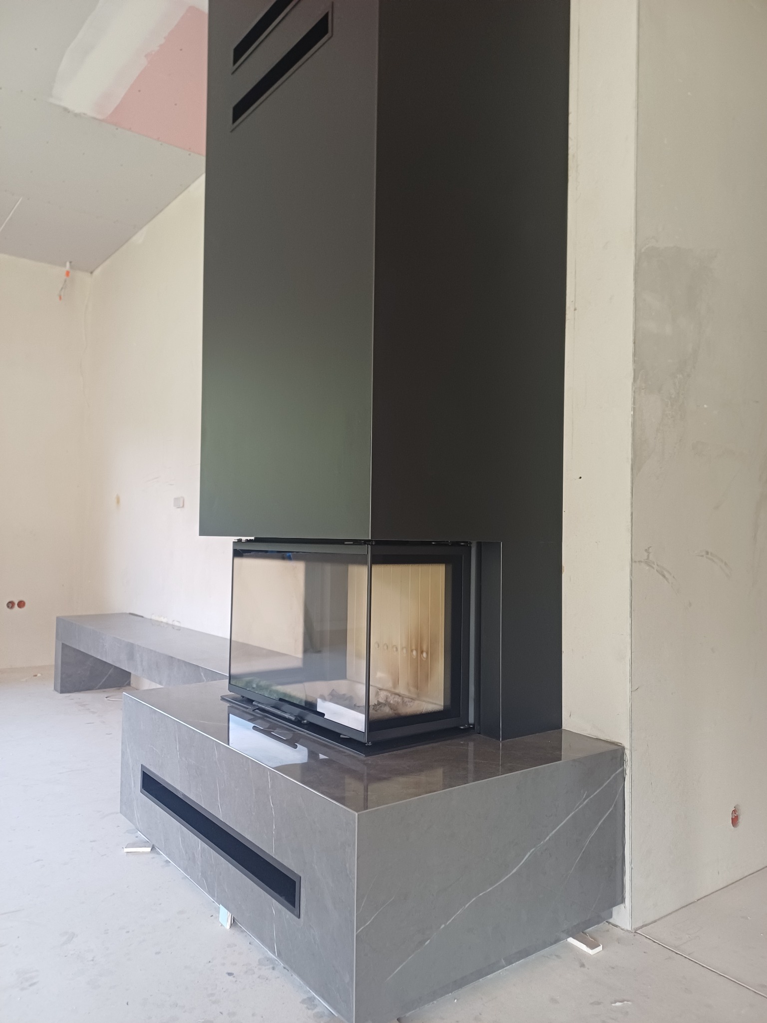 Nowoczesny kominek z szarą obudową imitującą marmur, w surowym wnętrzu. Widoczne niedokończone ściany i podłoga, co sugeruje etap wykończenia.