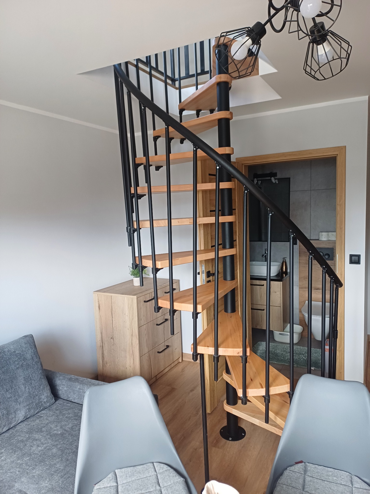 Spiralne schody z drewnianymi stopniami i czarną metalową konstrukcją w jasnym wnętrzu domu w Jastrzębiu. Widok na łazienkę w tle.
