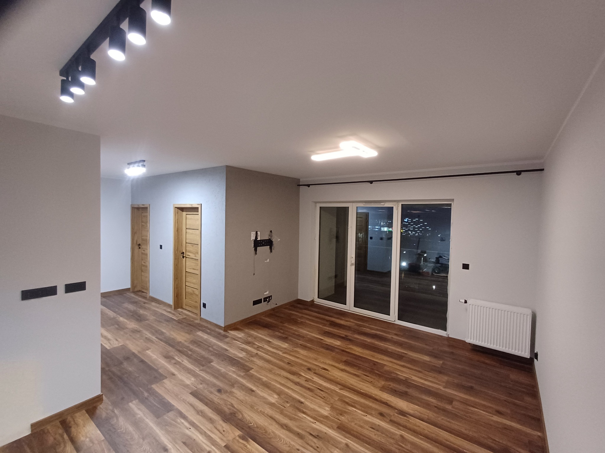 Wykończone wnętrze z drewnianą podłogą, białymi ścianami i nowoczesnym oświetleniem. Widoczne drzwi z drewnianą okleiną i duże okno balkonowe.