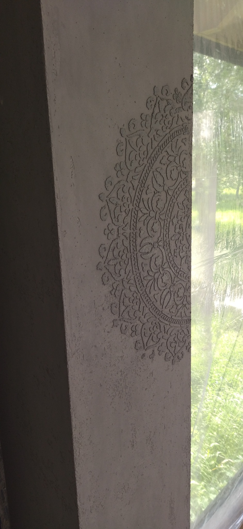 Dekoracyjna ściana z ornamentem w stylu mandali, wykonana z szarego tynku strukturalnego, częściowo zasłonięta folią ochronną przy oknie.