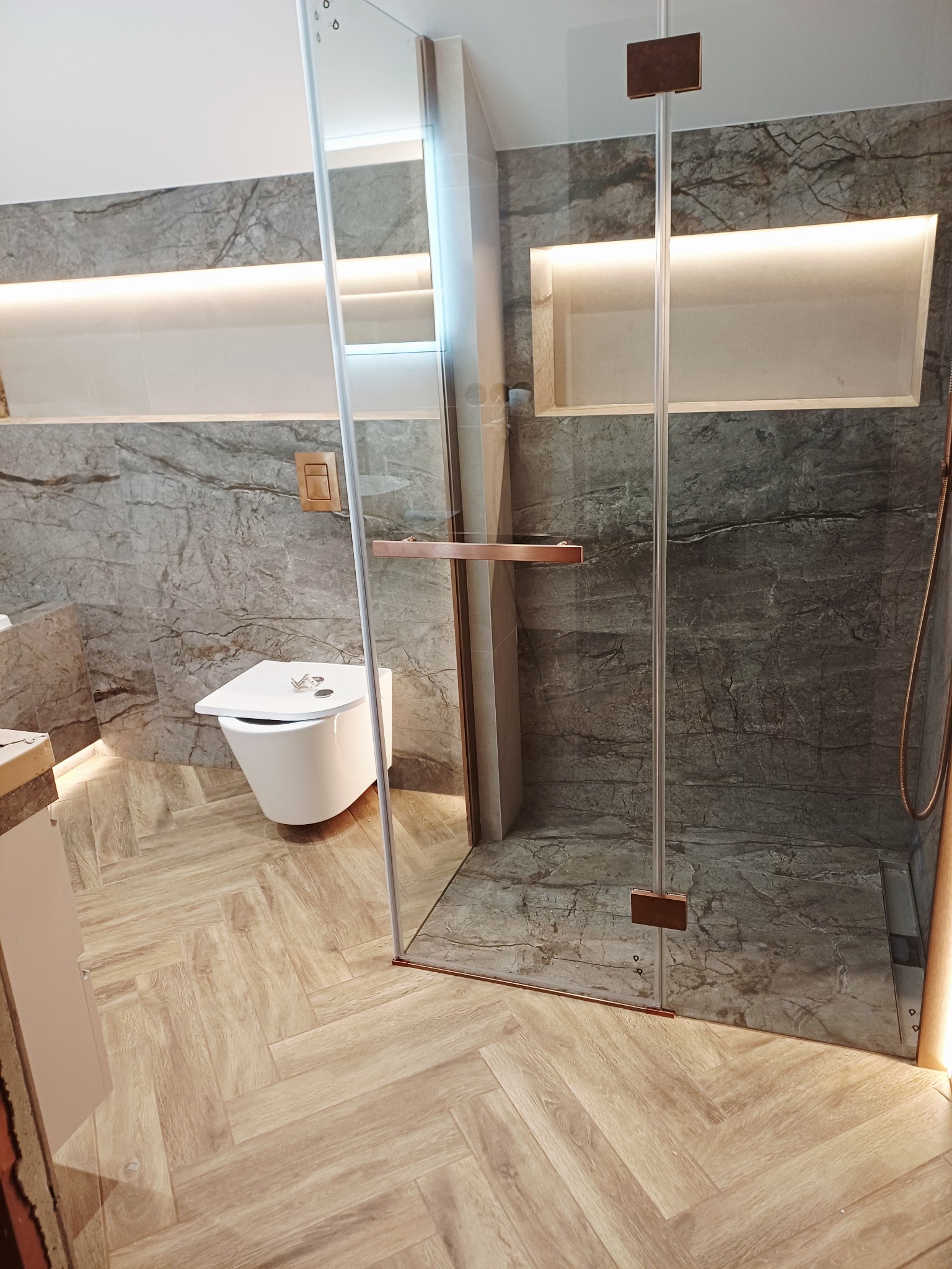 Nowoczesna łazienka z kabiną prysznicową typu walk-in, marmurowymi płytkami i drewnopodobną podłogą ułożoną w jodełkę. W tle podświetlane wnęki.